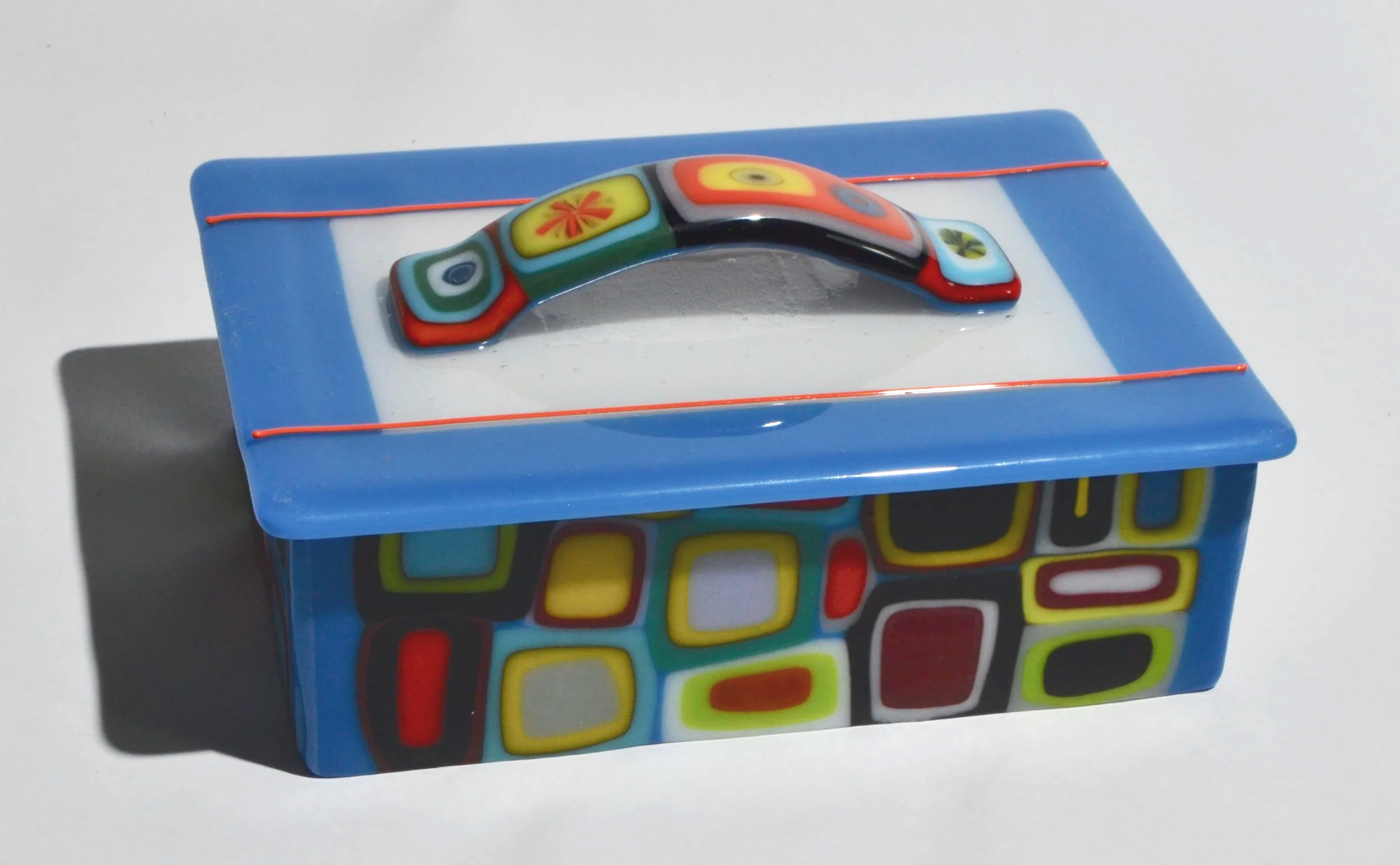 Colorful decorative box with multicolored geometric patterns and a curved glass handle on top, painted in bright colors including blue, red, yellow, and black.