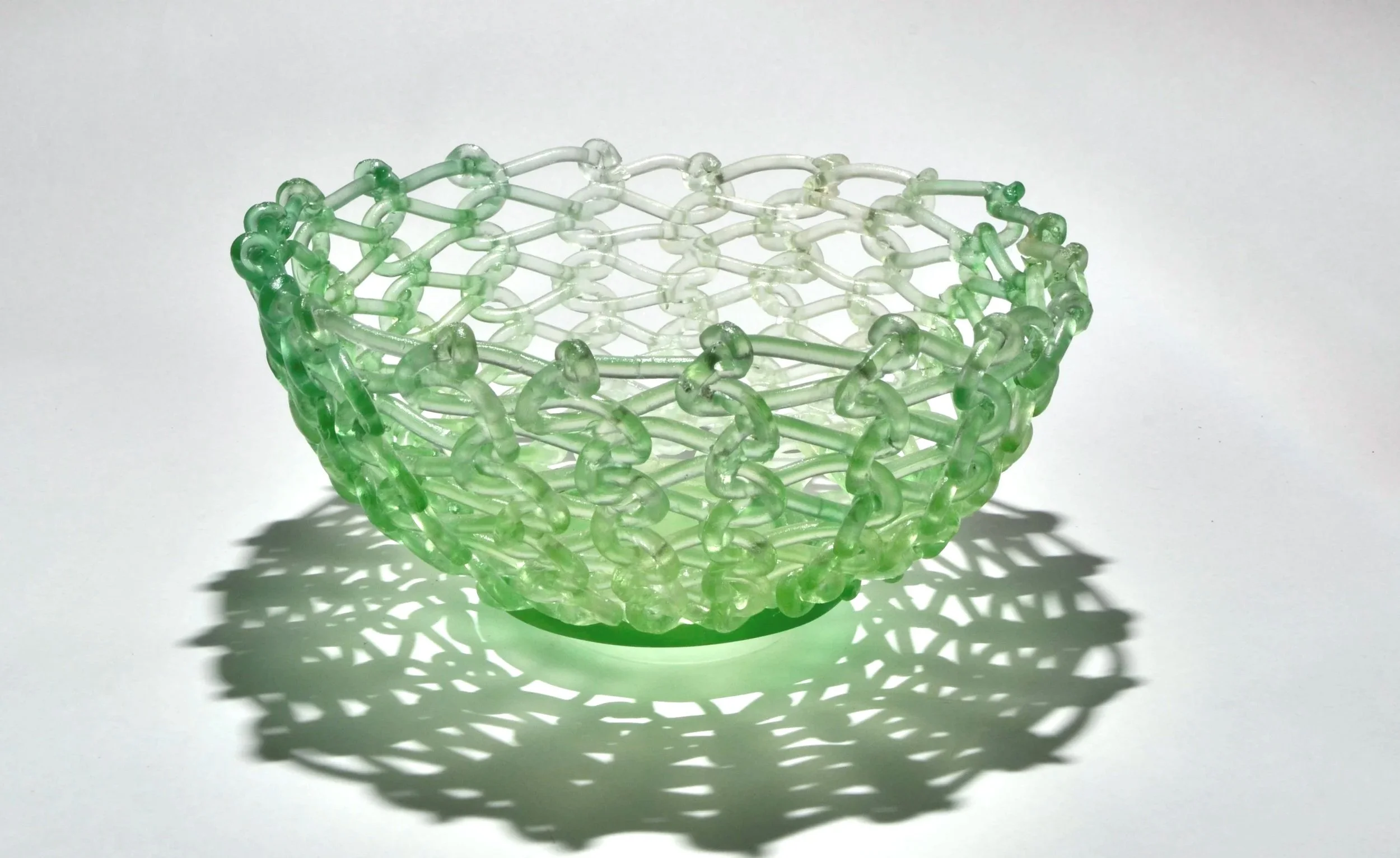 A green and clear plastic basket with a lattice design, casting a patterned shadow on a white background.