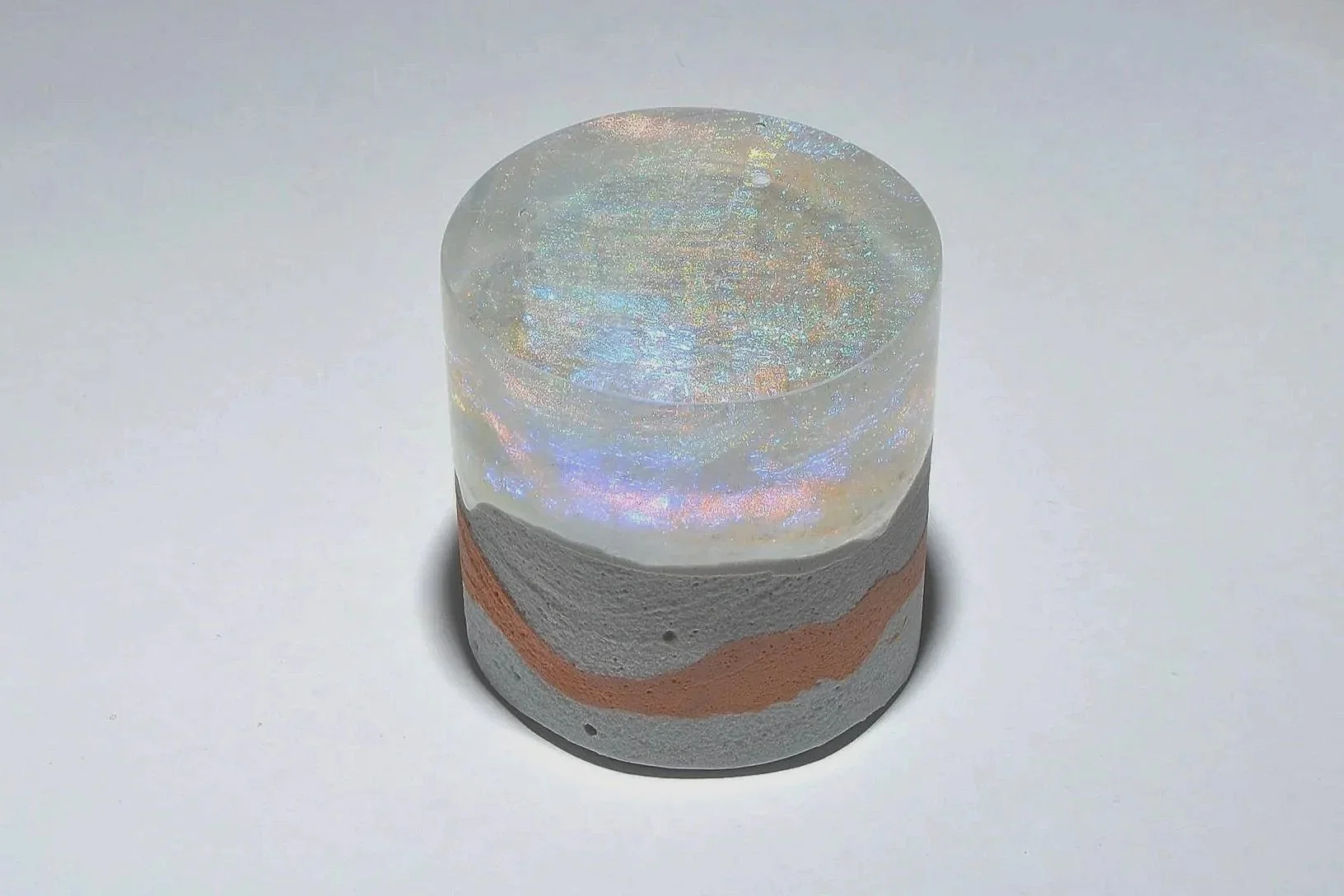 A small, cylindrical object with a base made of layered gray and pinkish clay-like material, topped with a clear, holographic, iridescent resin or glass cap.