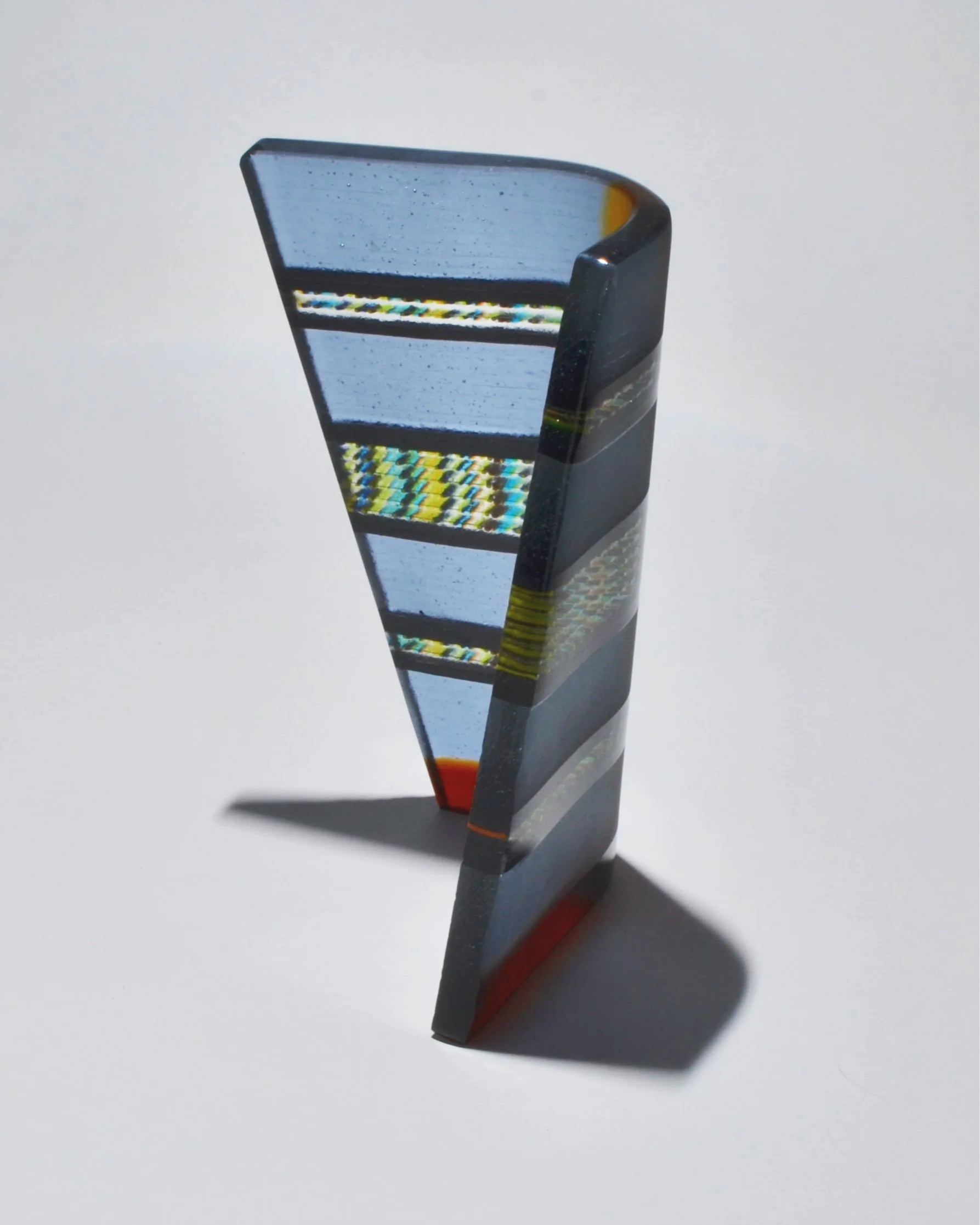 Close-up of a colorful, abstract glass sculpture with geometric patterns, casting a shadow on a plain white surface.