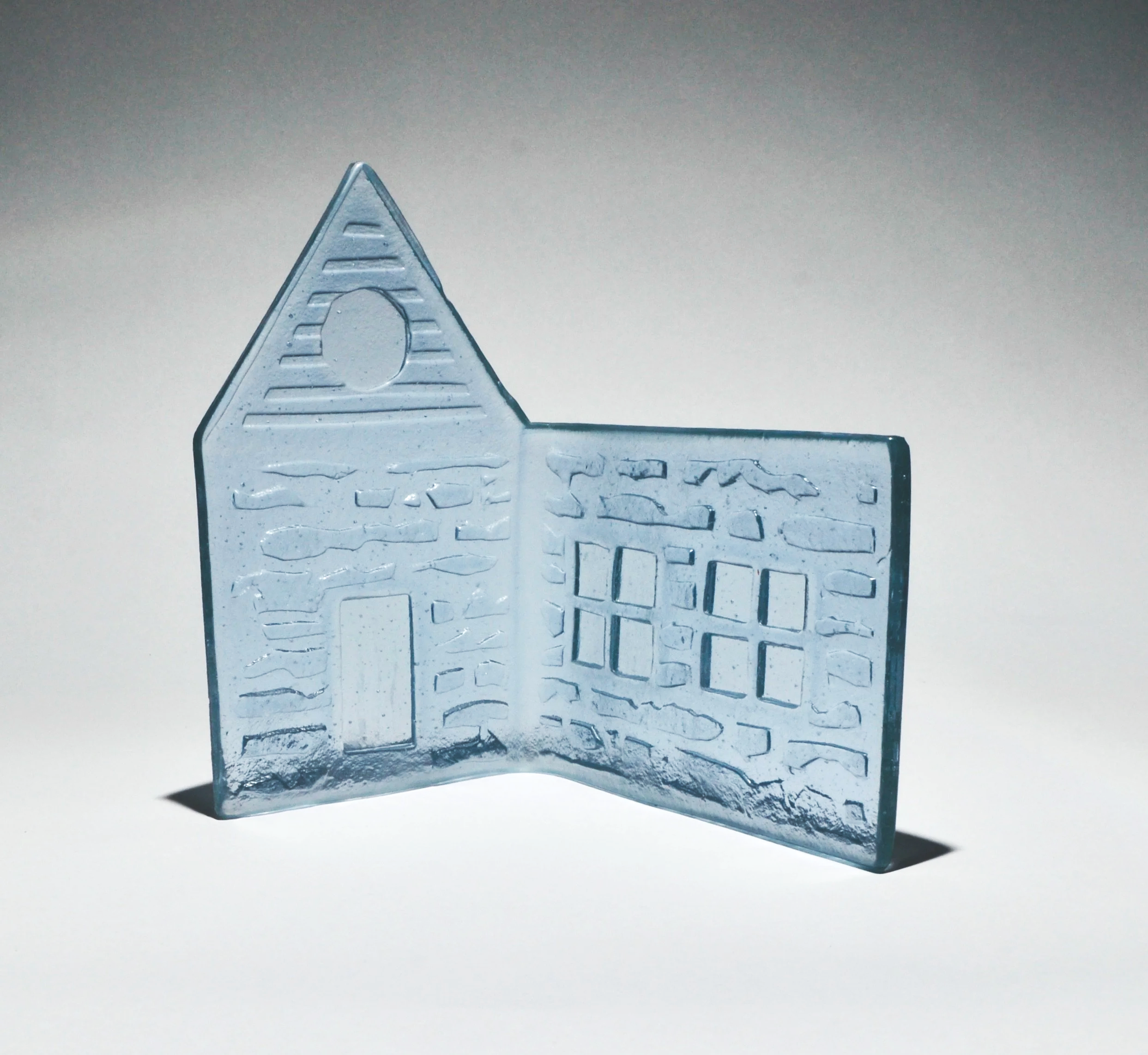 Glass house-shaped paperweight with etched window and door details, casting a shadow on a plain background.