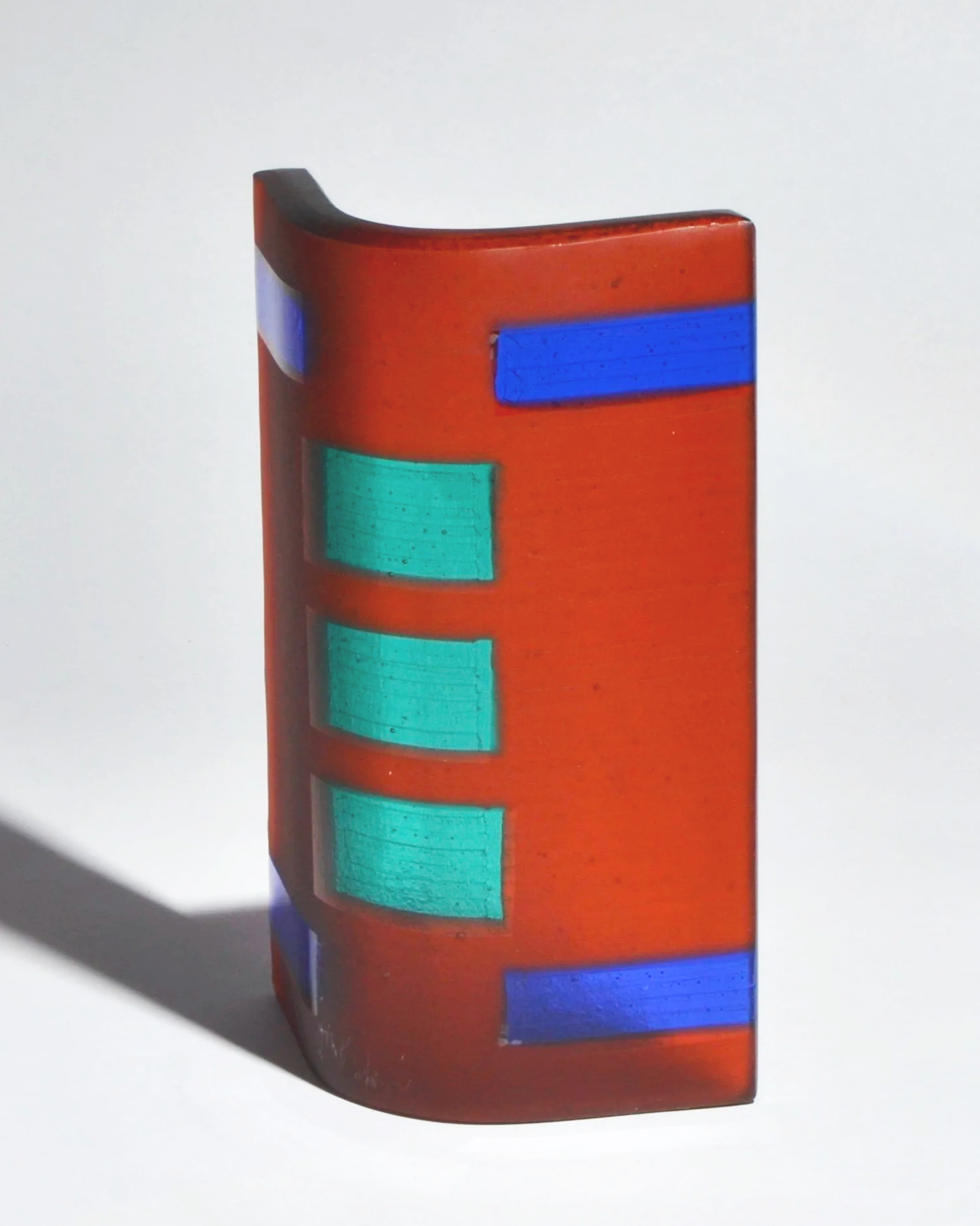 A colorful glass sculpture with an orange base, featuring blue and green rectangular accents, casting a shadow on a white background.