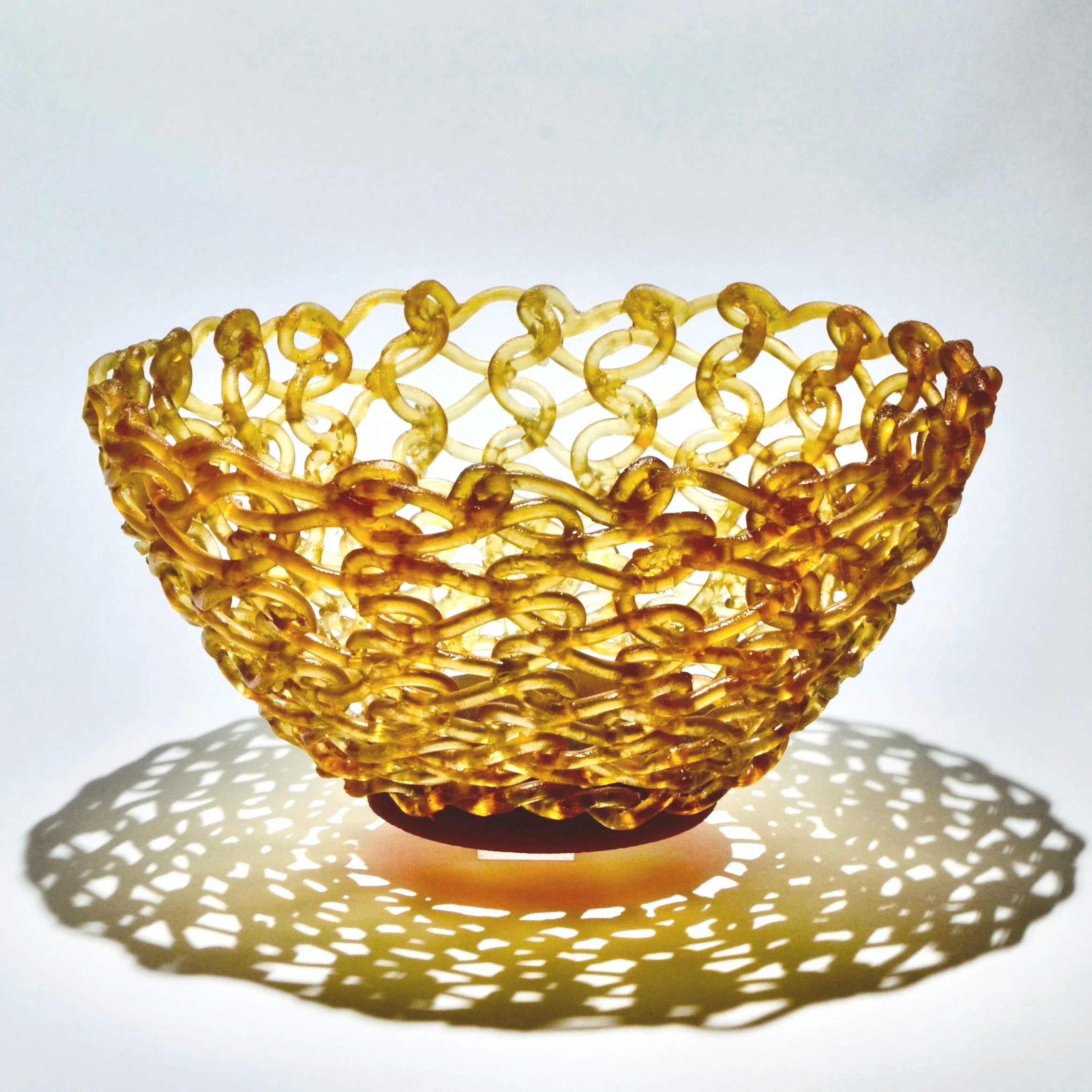 A decorative, open-weave, gold-colored bowl with intricate chain-link pattern, casting a patterned shadow on the white surface below.