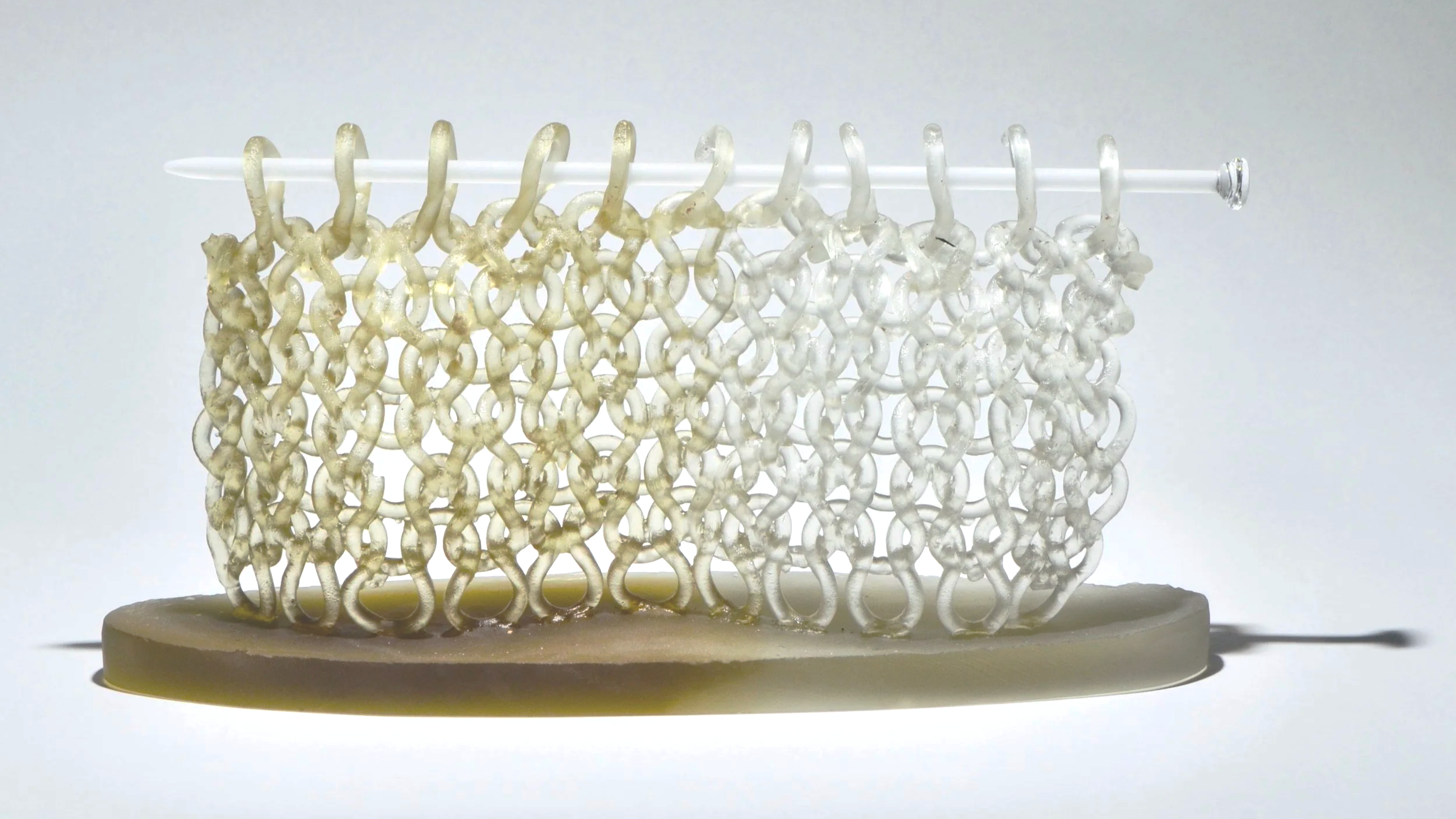 Close-up image of a small 3D-printed structure made of intertwined loops, supported by a thin horizontal rod placed on an oval base, with a plain white background.