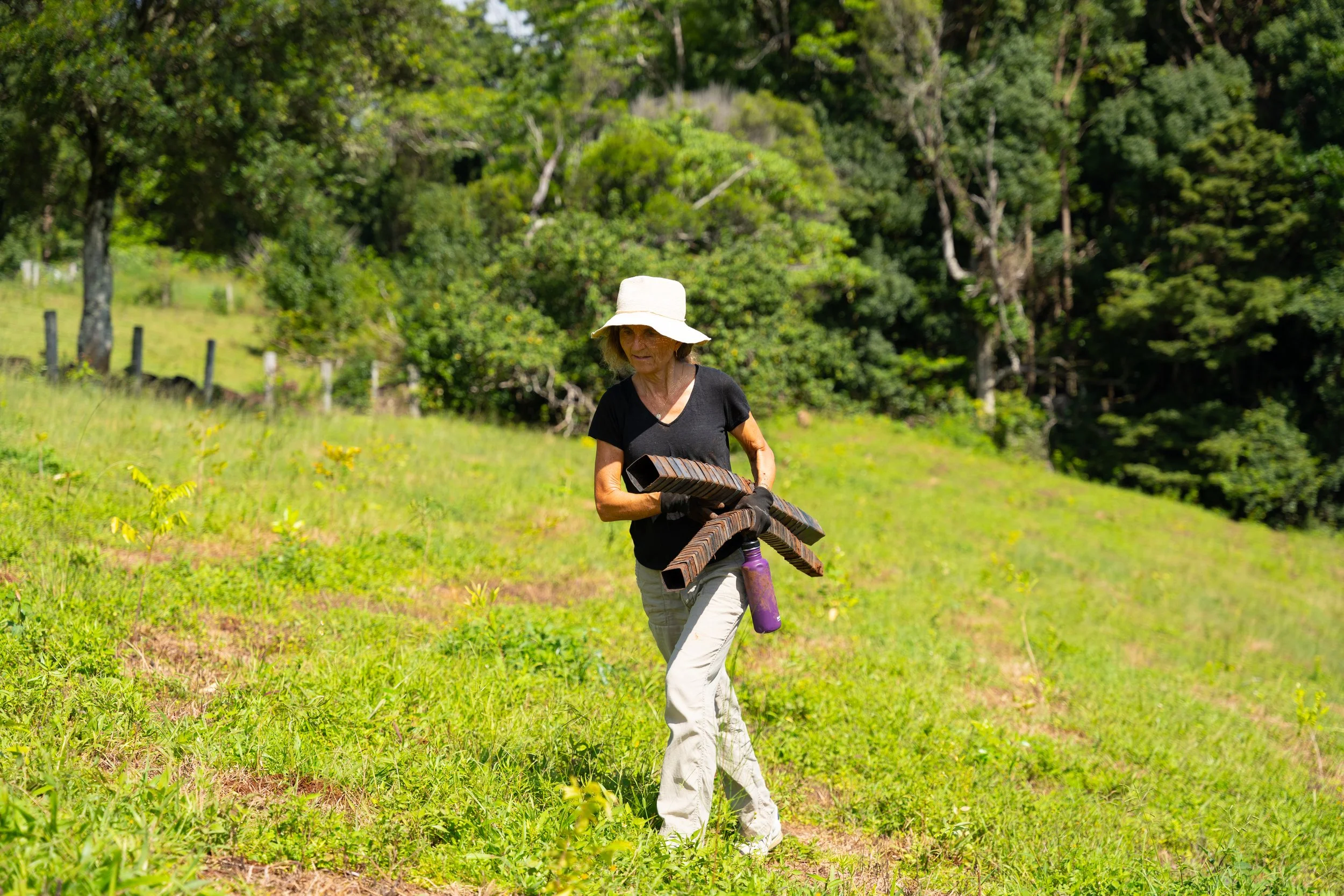 A woman in a white hat, black shirt, and beige pants carrying foam pool noodles and a water bottle in a green grassy field with trees in the background.