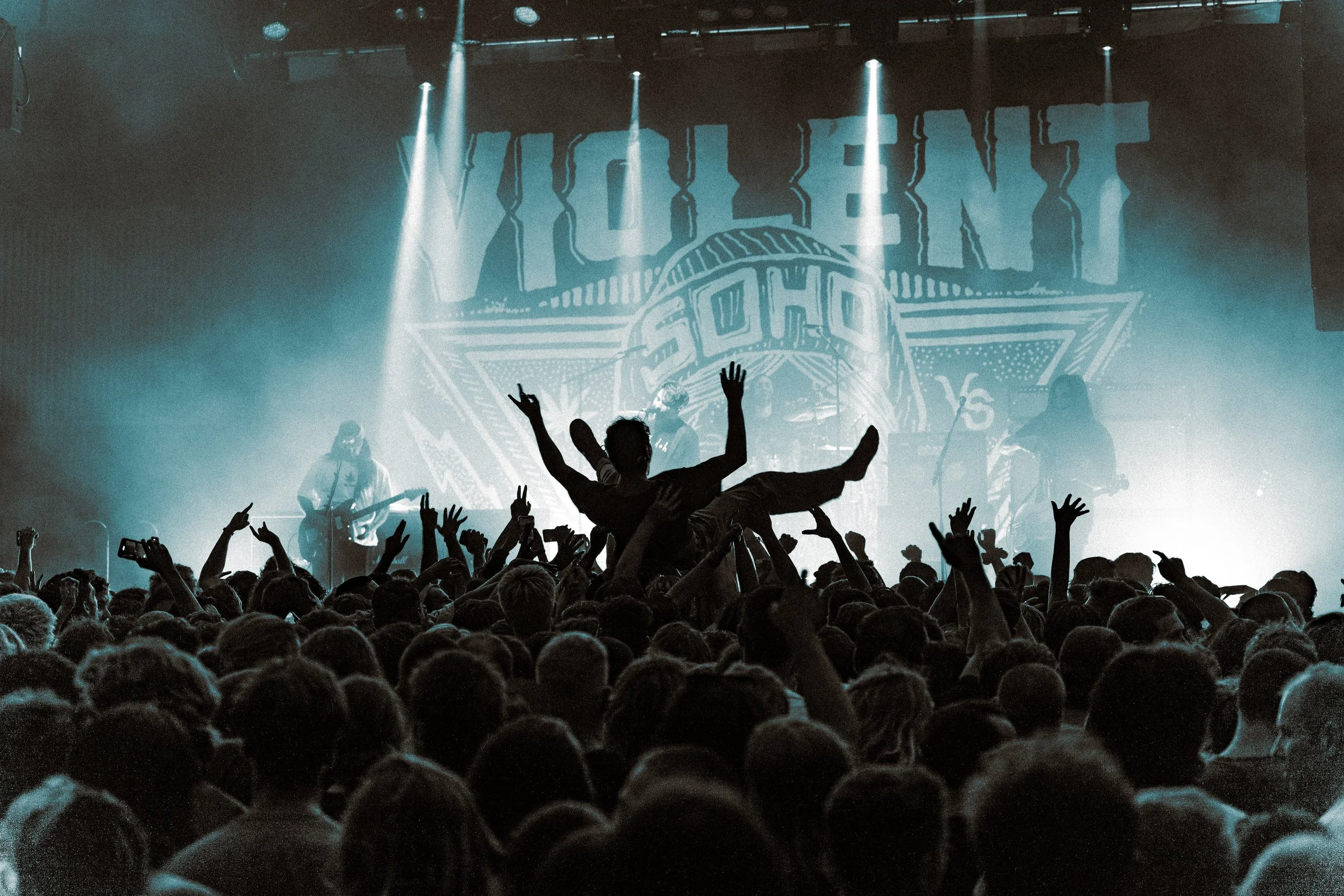 Crowd at the concert with a person being lifted on others' hands, performing on stage with band members, with the stage backdrop displaying the word "Violent".