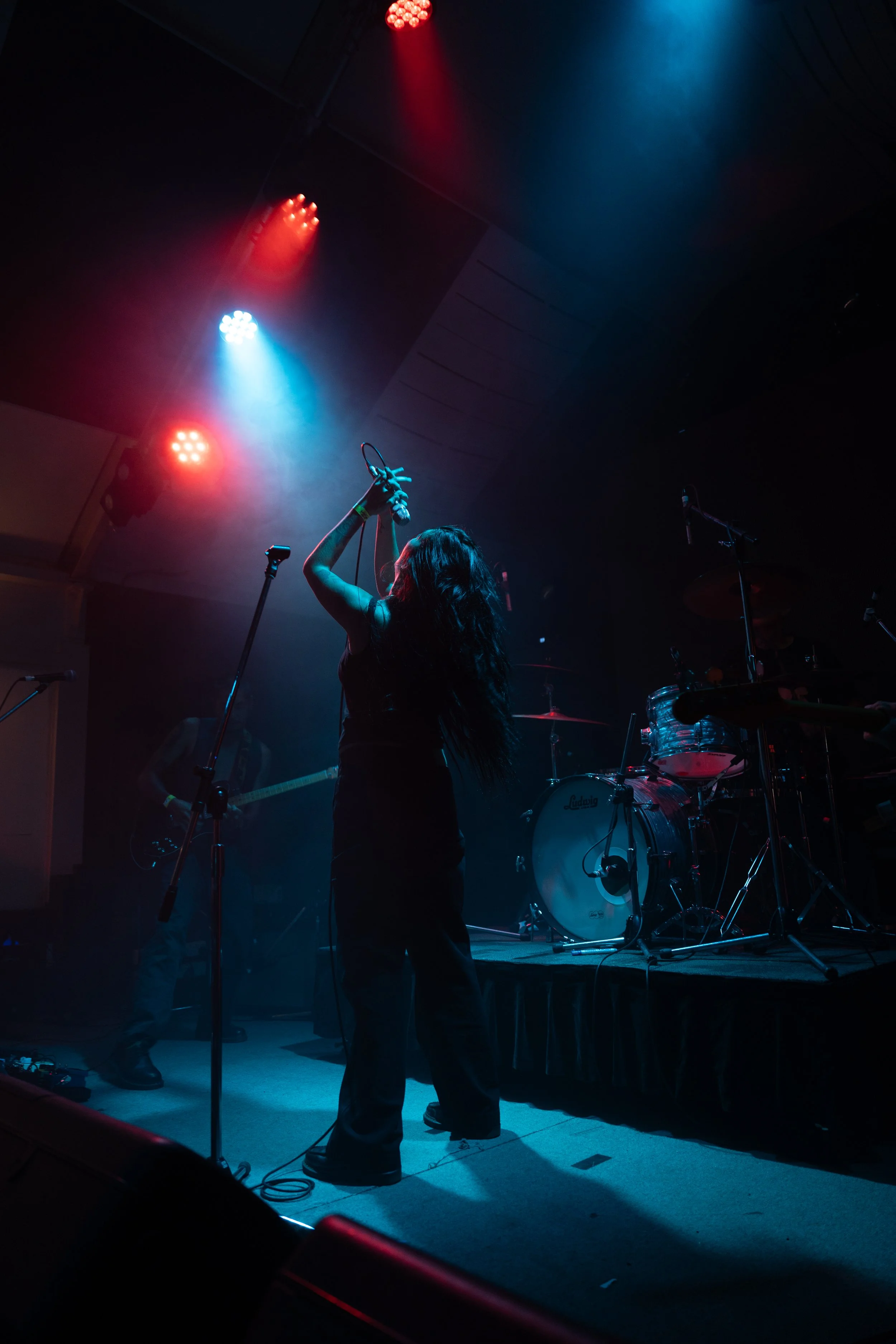 A female singer with long dark hair performing on stage at a concert, illuminated by red and blue stage lights, with a drummer and guitarist visible in the background.