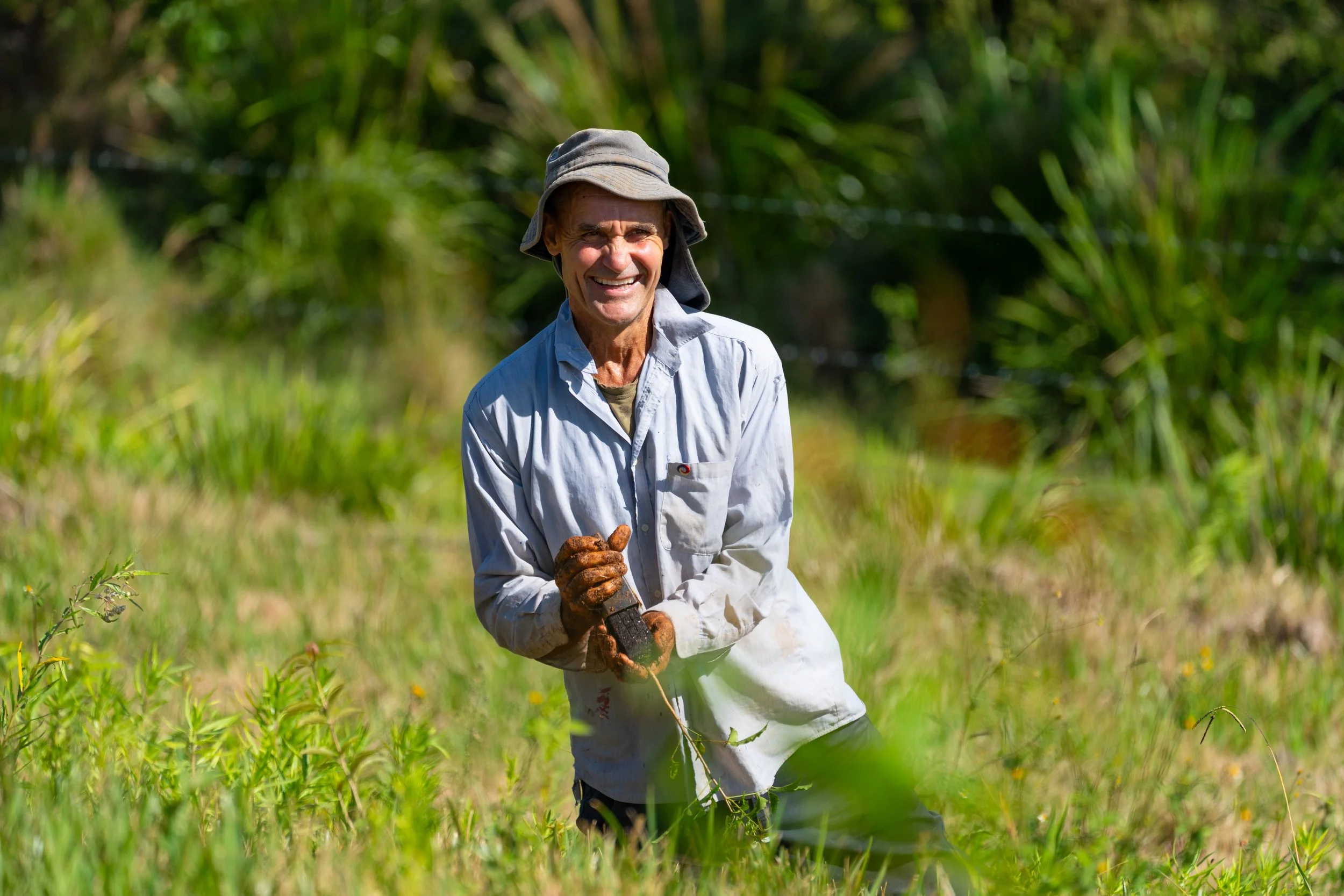 A smiling man in outdoor clothing and a wide-brimmed hat standing in a grassy field surrounded by greenery.