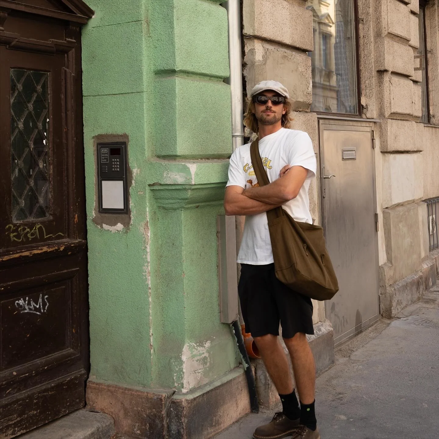 A man wearing sunglasses, a white cap, a white t-shirt, black shorts, and brown shoes stands with arms crossed on a city sidewalk next to a green building with a brown door and graffiti. He has a brown shoulder bag.