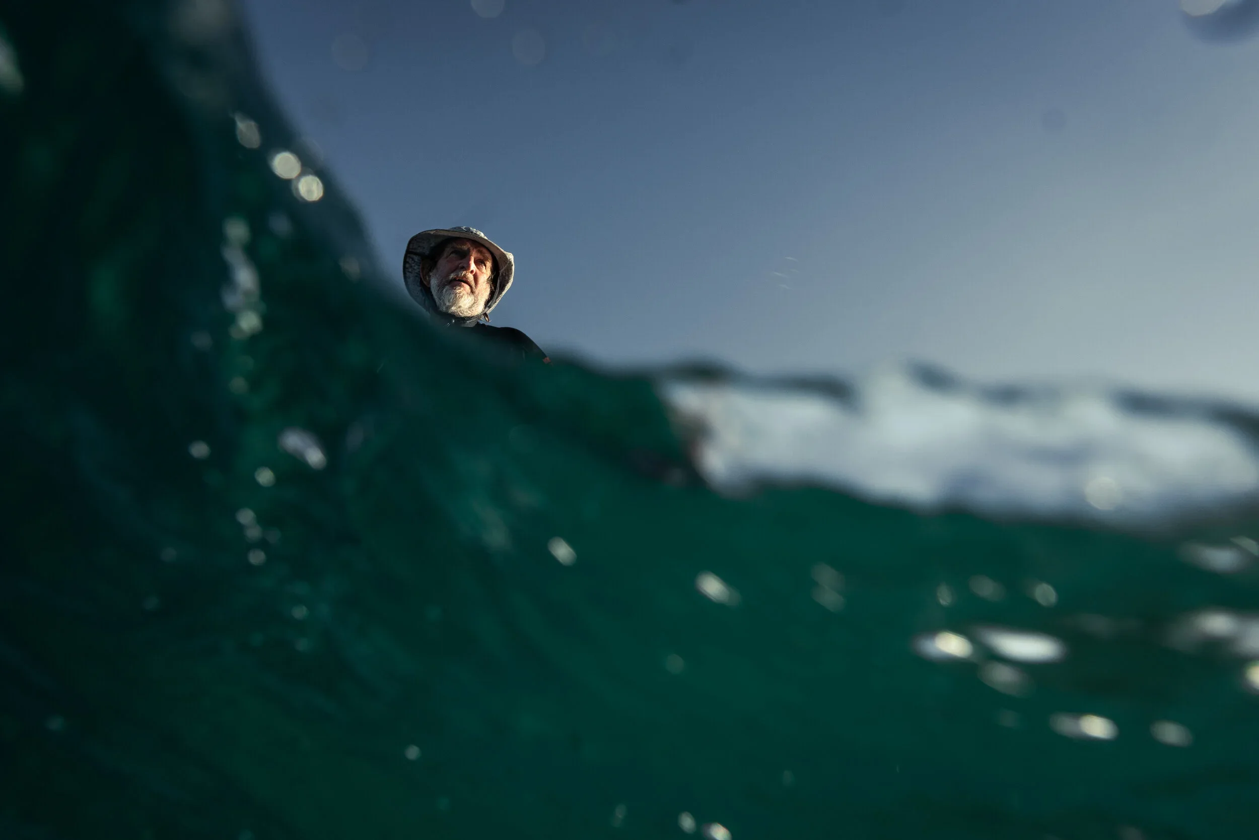 A person with a beard wearing a wide-brimmed hat looks over a wave in the ocean on a clear day.