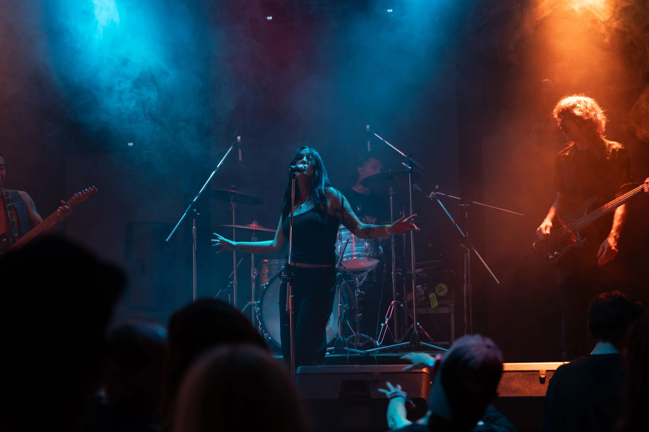 A band performing live on stage with colorful lighting, including a female singer in the center, a drummer behind her, and two guitarists on either side.