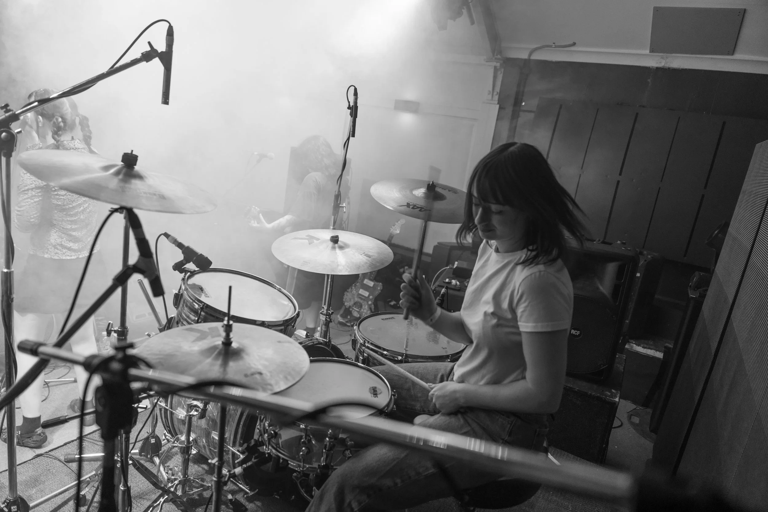 A woman playing drums in a recording studio with microphones and other band members in the background.