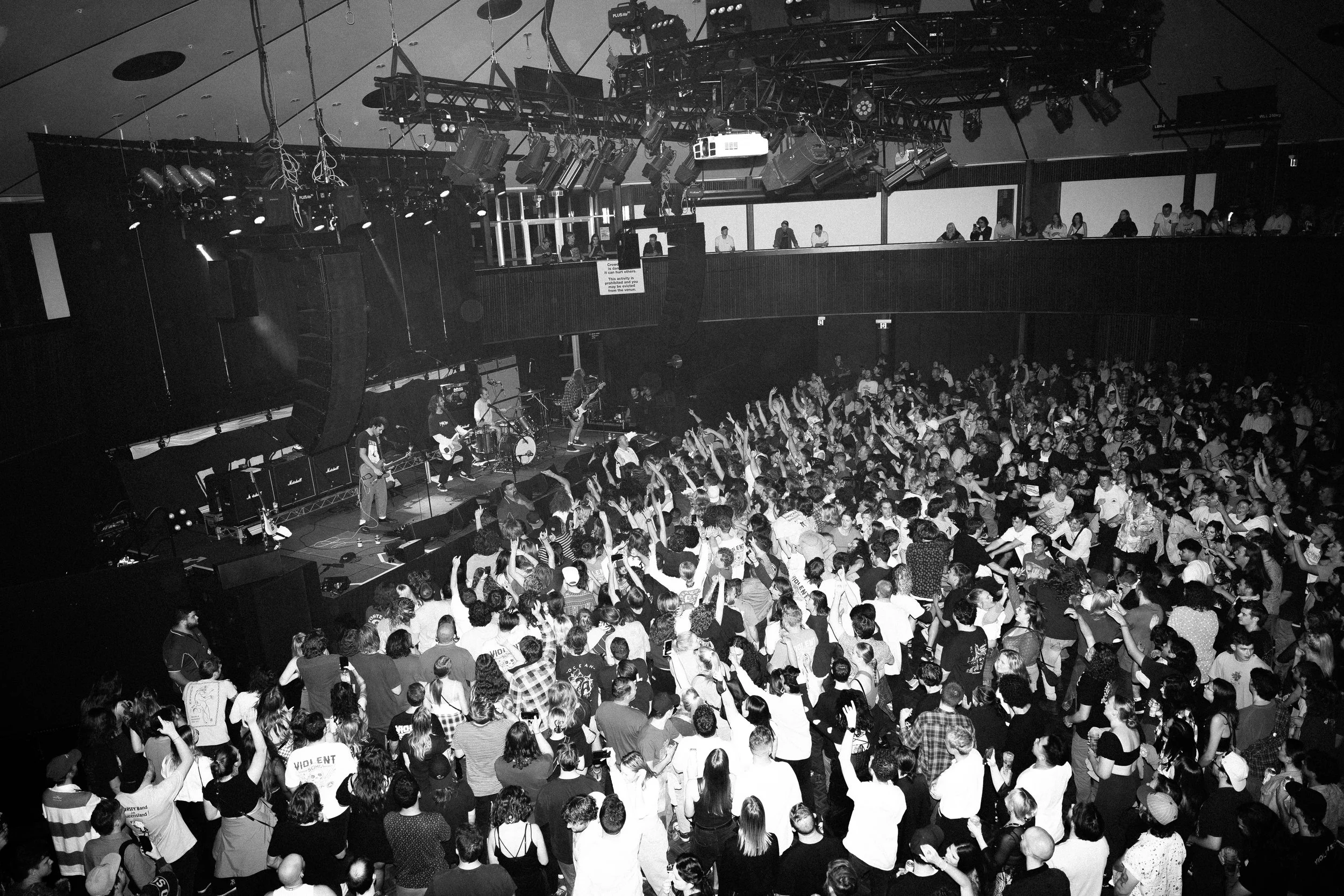 Black and white photo of a crowded concert with a band performing on stage, multiple audience members in front dancing or raising their arms, and balcony with additional people watching.