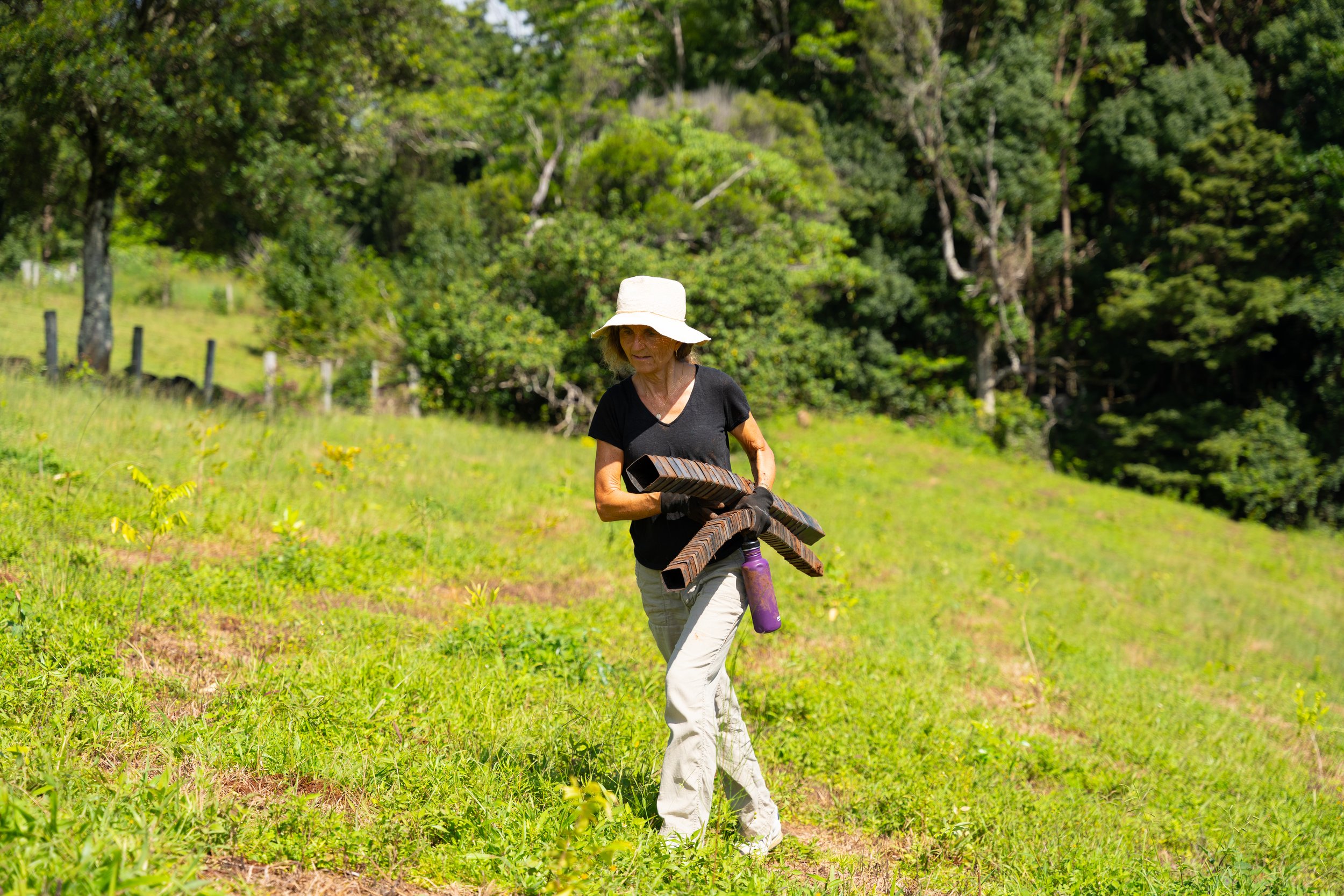 Woman wearing a sun hat and black shirt carrying pieces of garden pipes and a water bottle through a green grassy field with trees in the background.