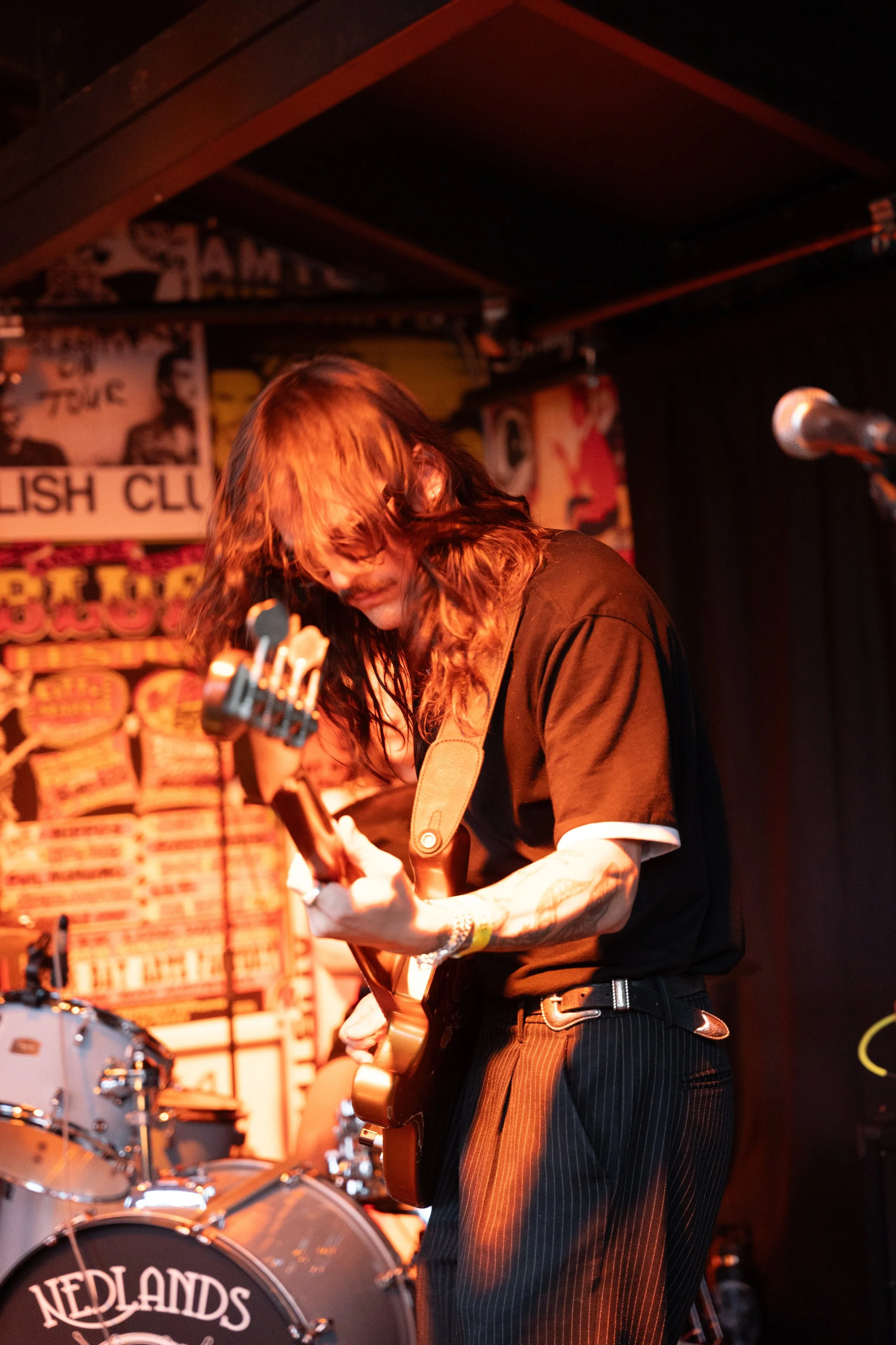 A male musician with long hair and a mustache playing an electric guitar on stage at a music venue. There is a drum set behind him and posters on the wall in the background.