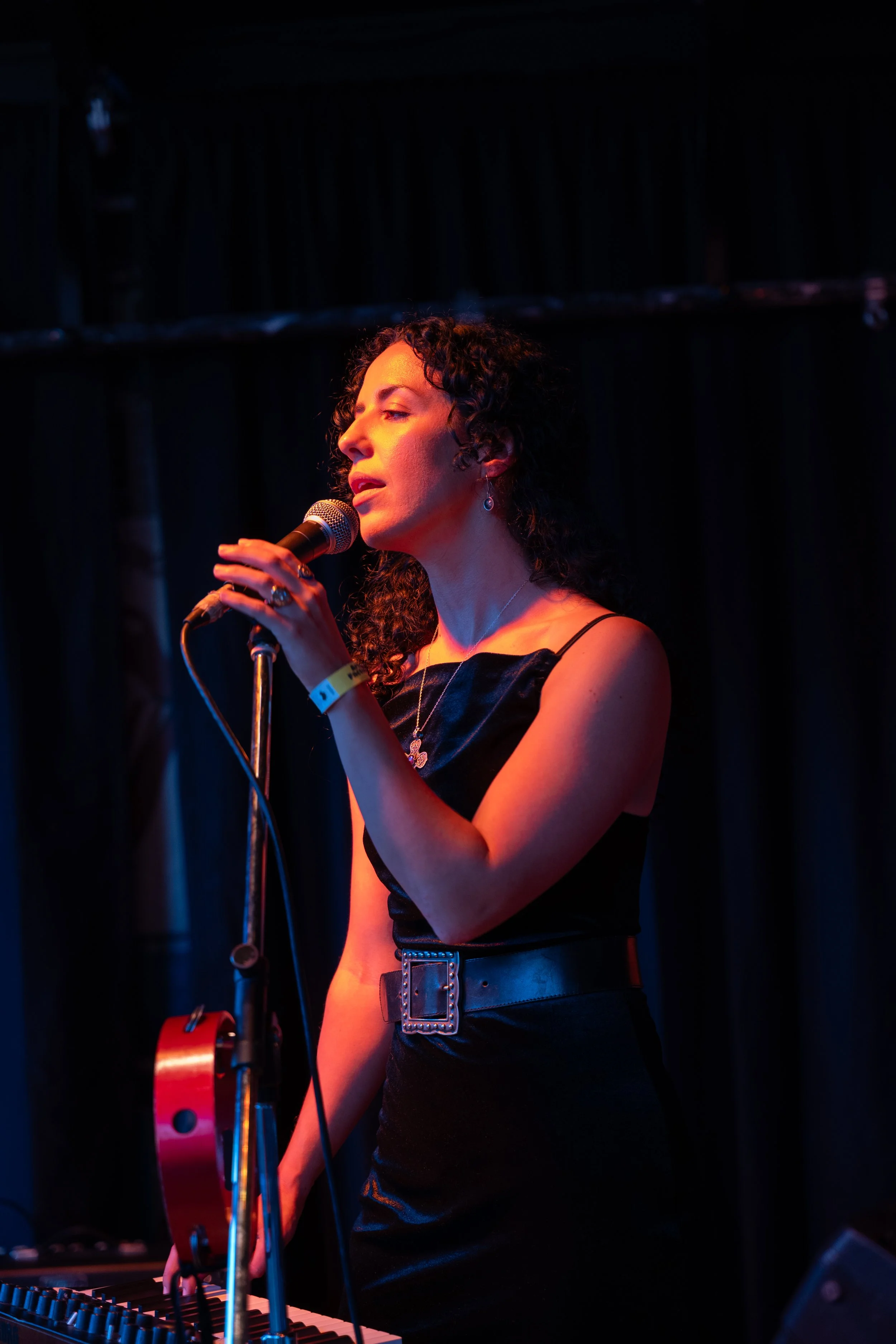 A woman with curly dark hair singing into a microphone on stage, with a keyboard in front of her and a dark curtain behind her.