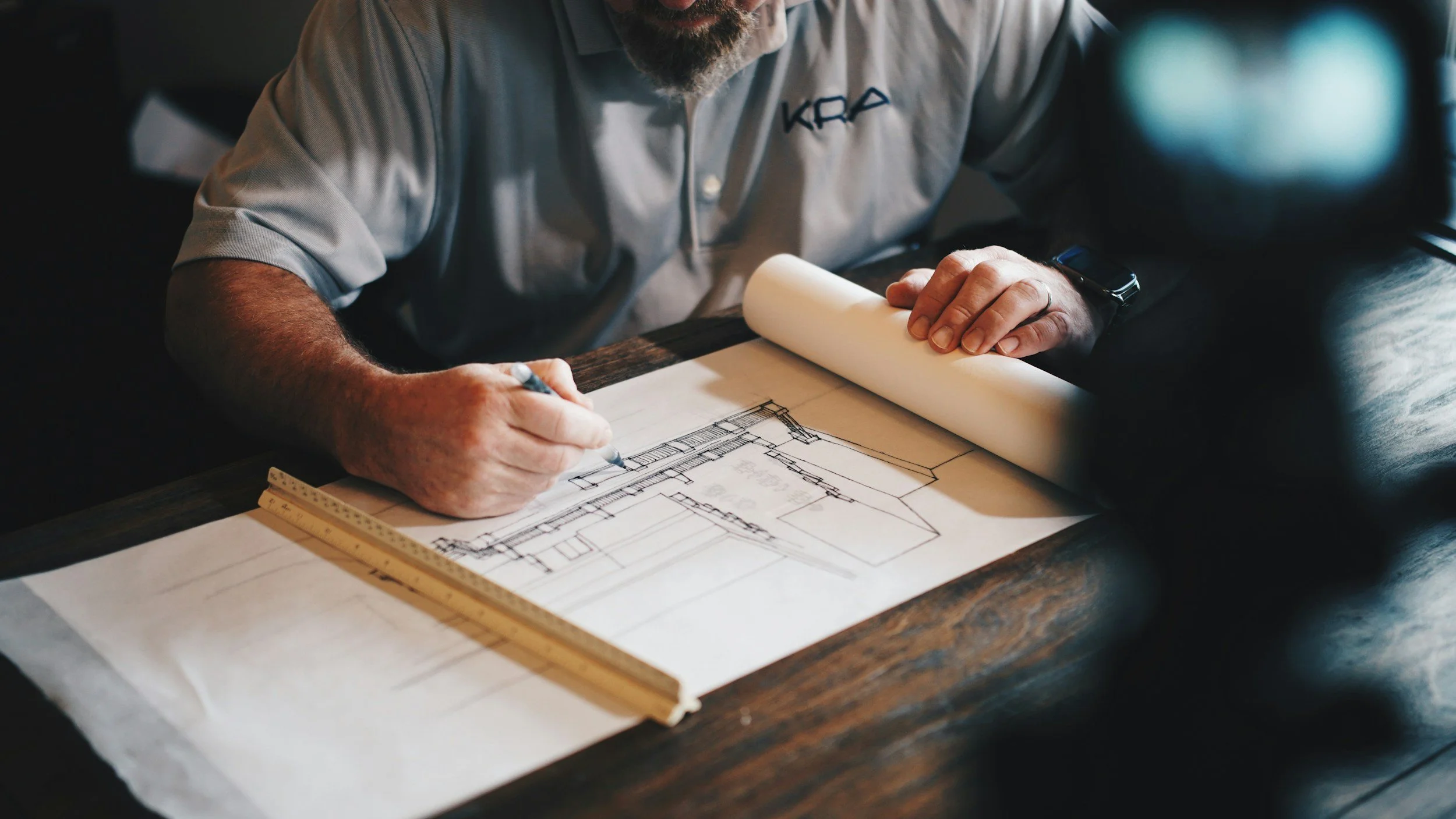 A man is sketching architectural blueprints on paper at a wooden table, with a ruler and rolled-up paper nearby.