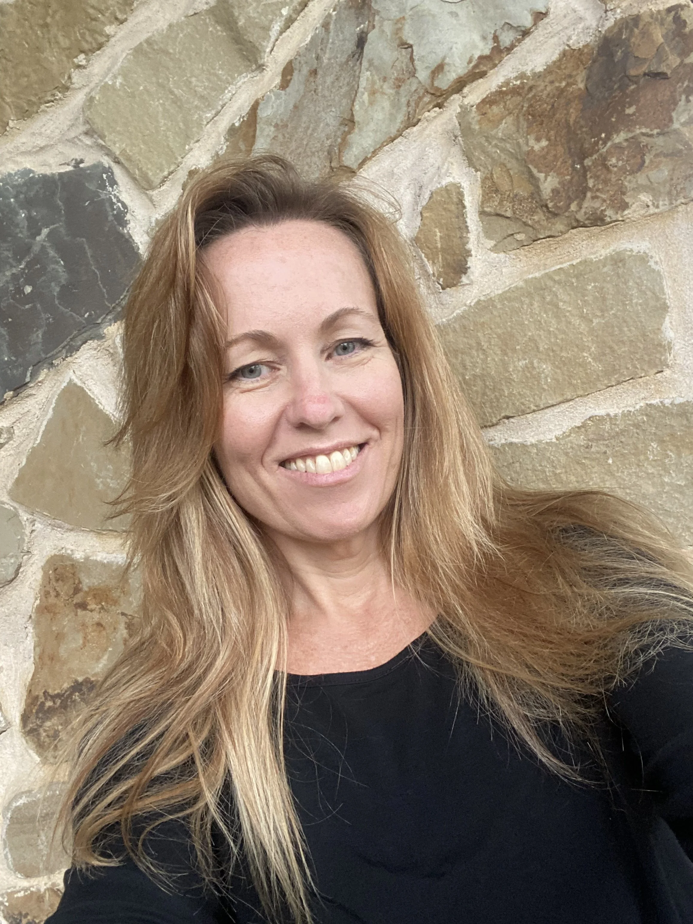 A woman with long, blonde hair wearing a black shirt, smiling, in front of a stone wall.