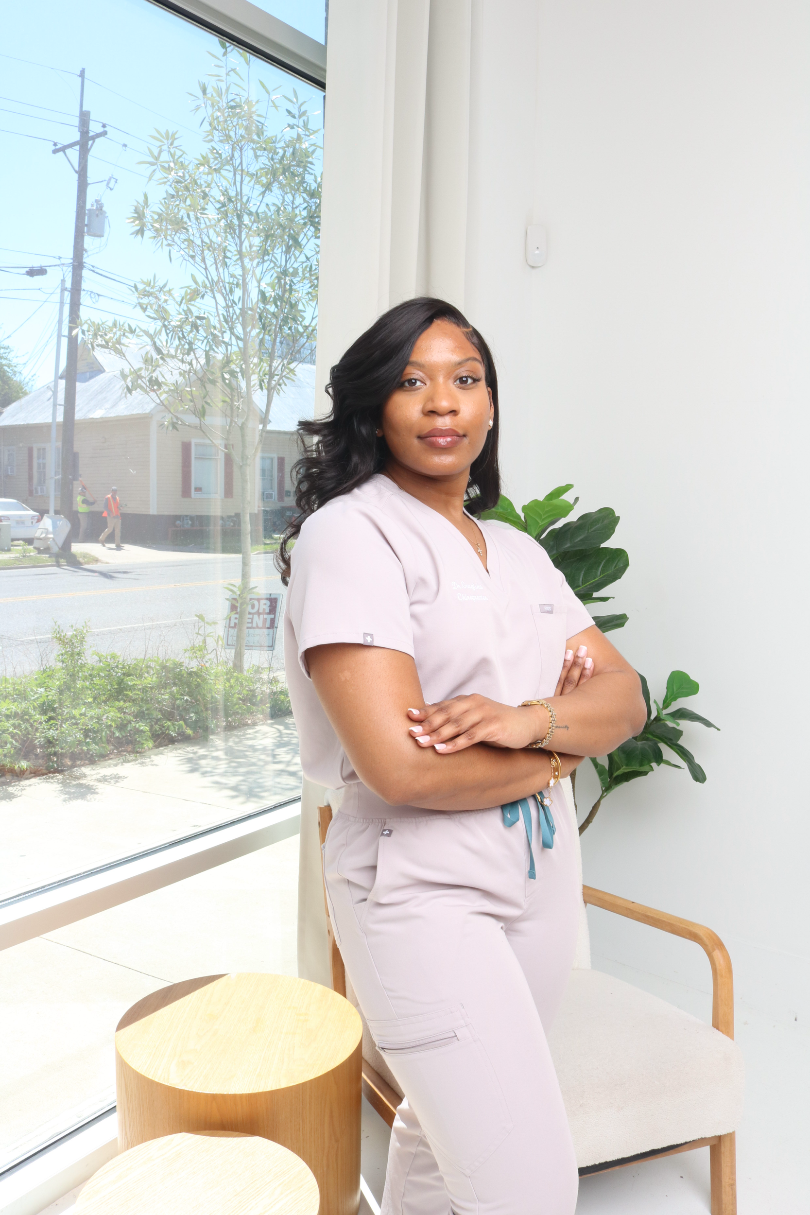 A woman in medical scrubs standing with arms crossed in a bright room with large window and indoor plant.