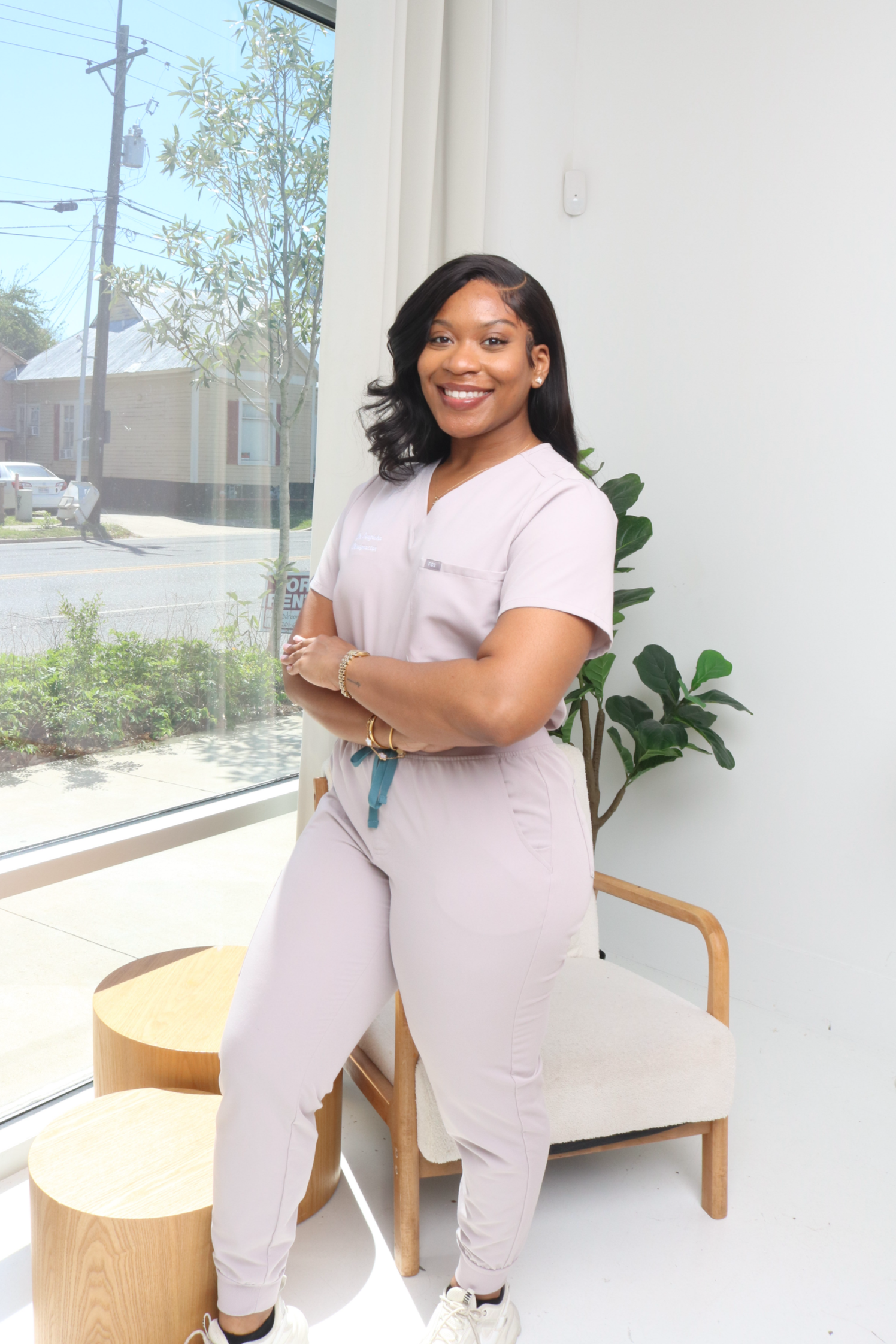 A young woman with dark hair and a bright smile, dressed in light medical scrubs, standing inside a room with a large window and a potted plant, crossing her arms.