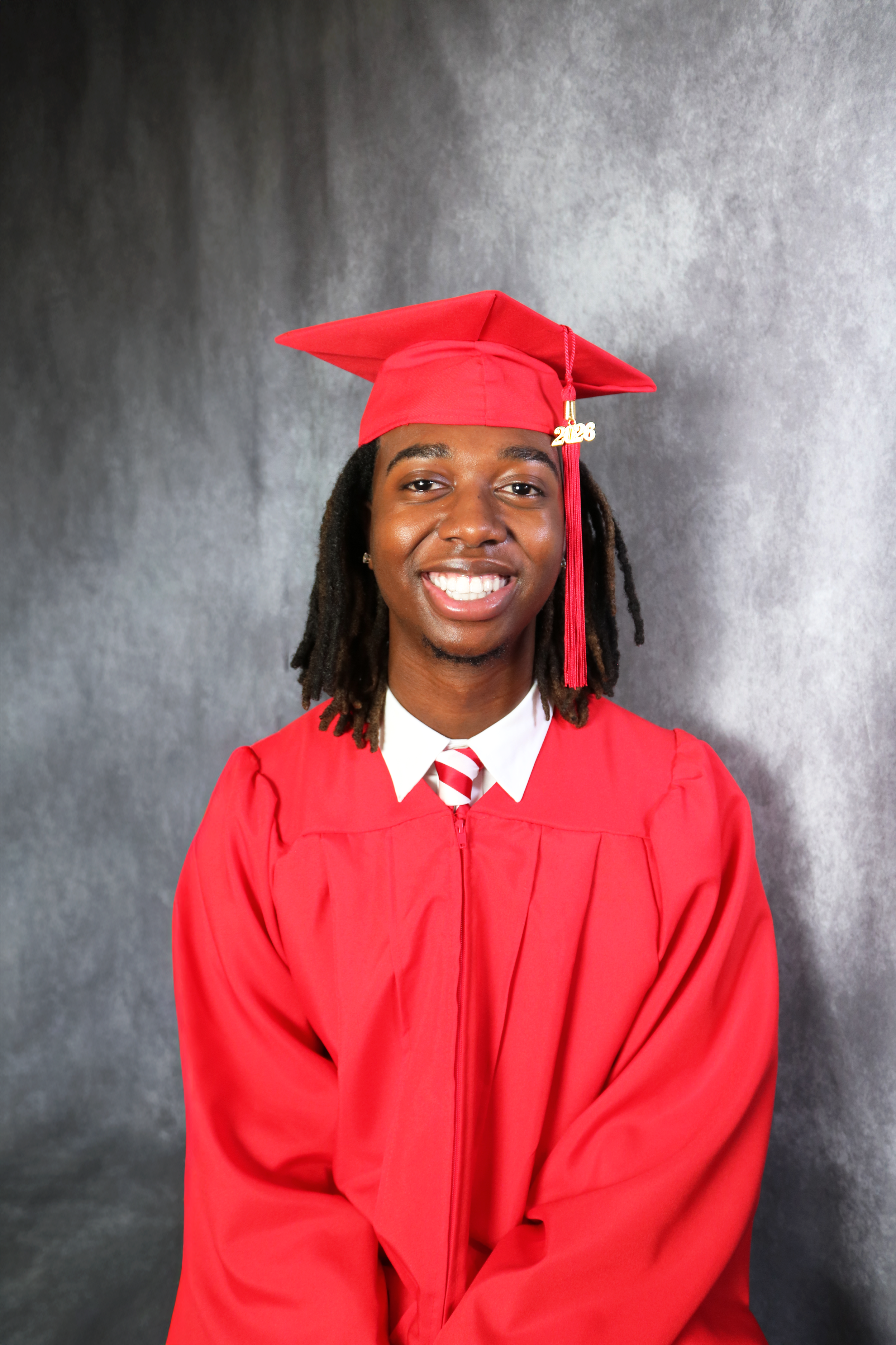 A young man wearing a red graduation cap and gown, smiling, posed against a plain gray background.