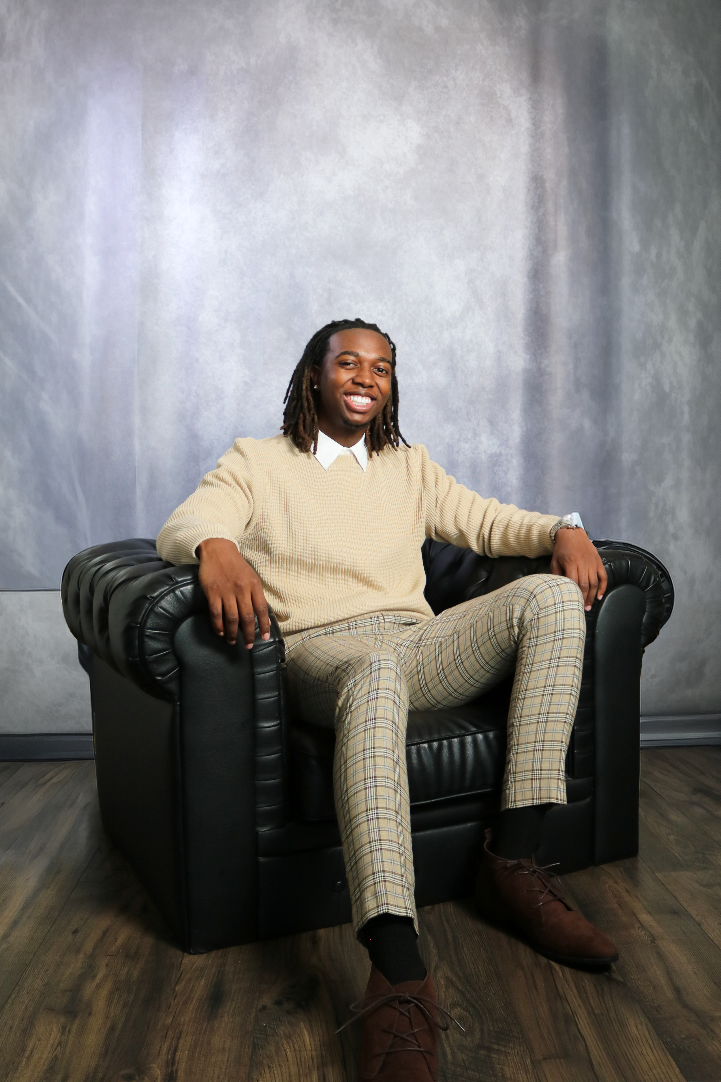 A man with dreadlocks sitting on a black leather armchair with a gray textured backdrop. He is smiling, wearing a beige sweater over a white collared shirt, plaid pants, and brown boots.