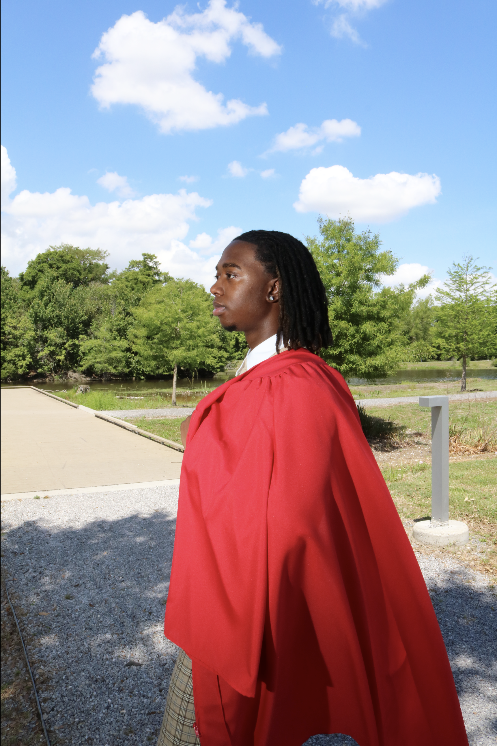 Profile of a young Black man with dreadlocks wearing a red graduation gown outdoors on a sunny day with green trees and a blue sky with white clouds.