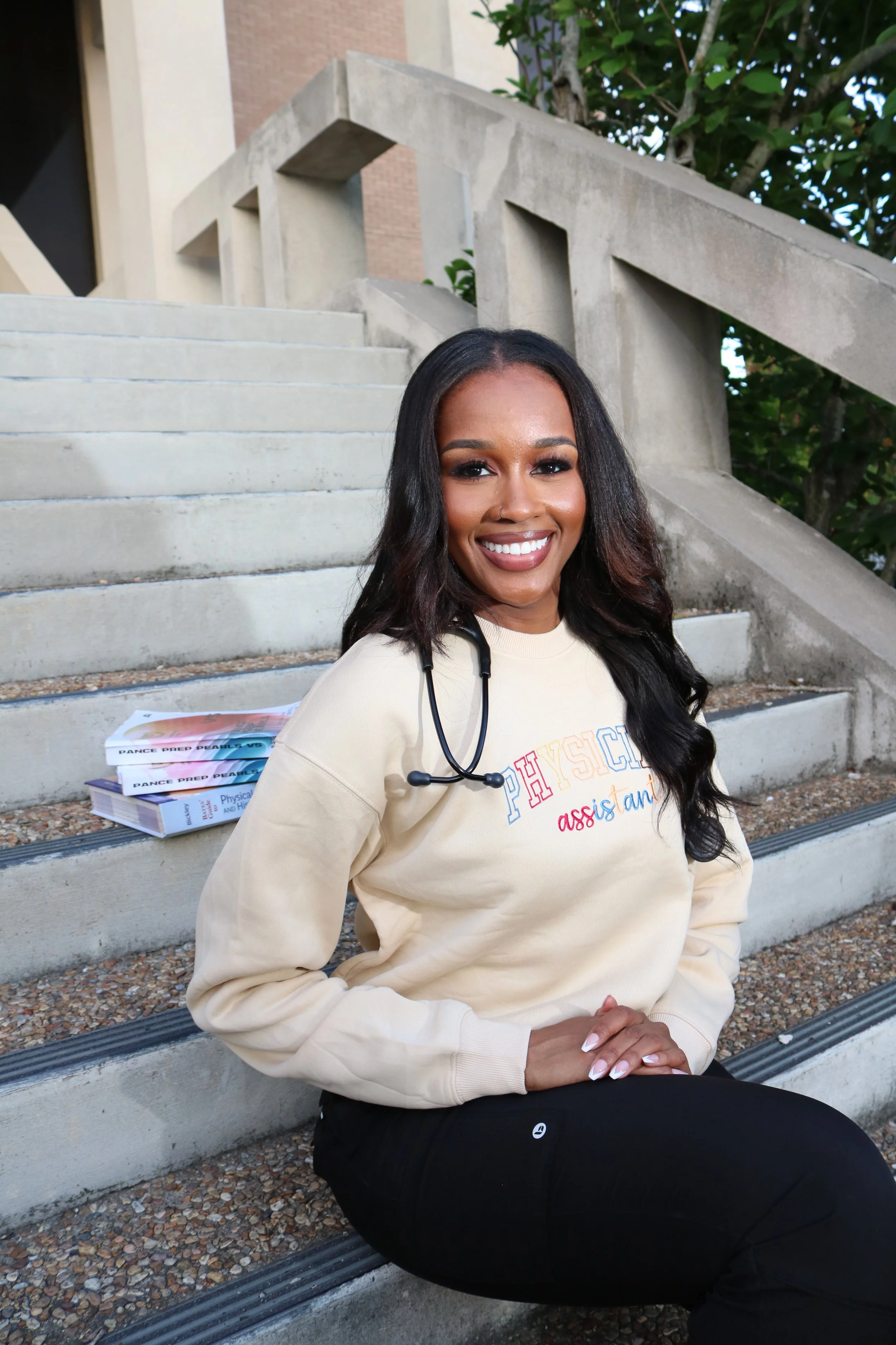 A smiling woman with dark hair and a nose ring, sitting on concrete steps outdoors next to a stack of books. She is wearing a cream-colored sweatshirt with colorful text and a stethoscope around her neck. There are green trees and a building in the b