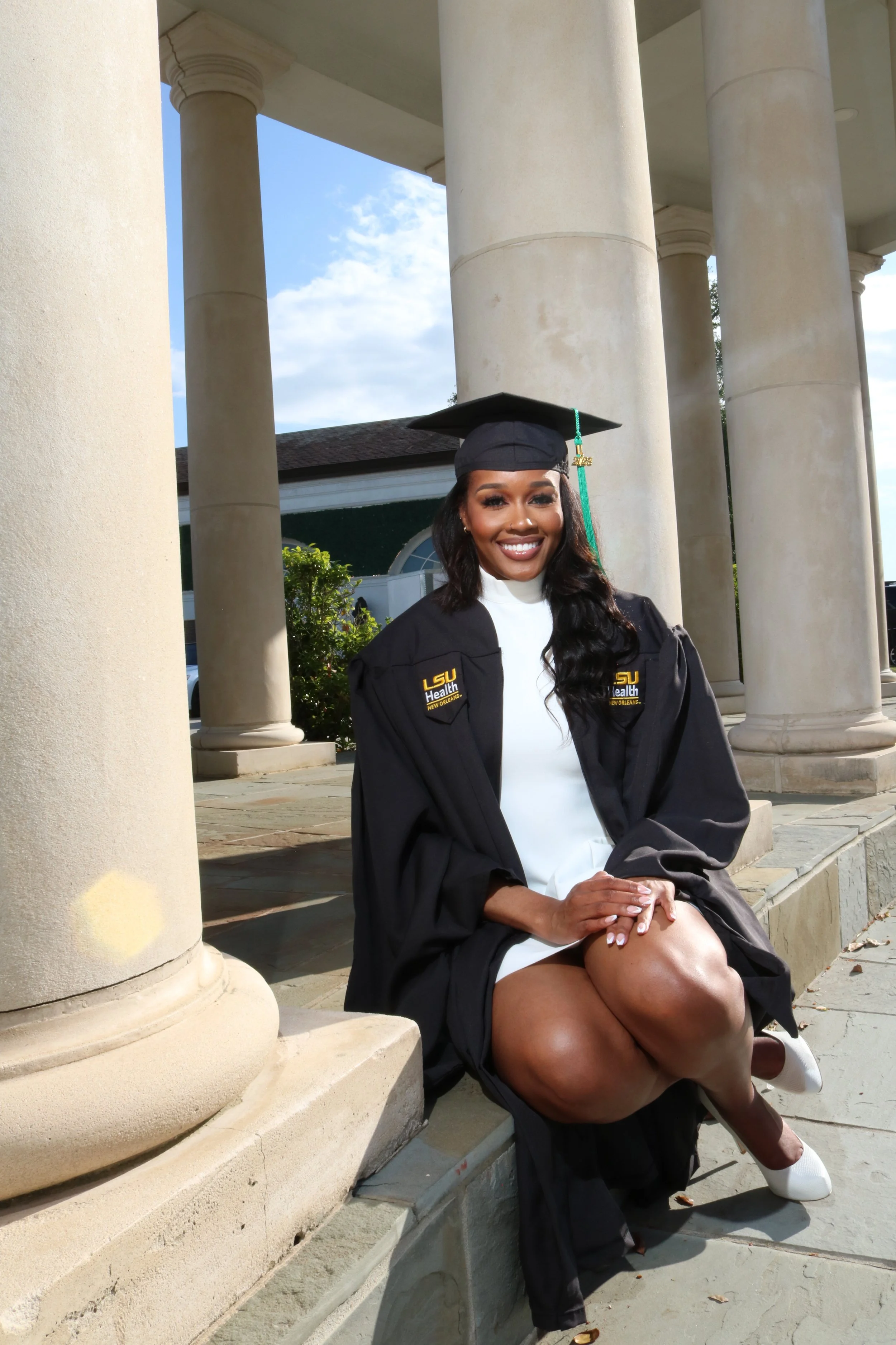 A young woman in a graduation cap and gown sitting on steps outdoors, smiling, with columns and a building in the background.