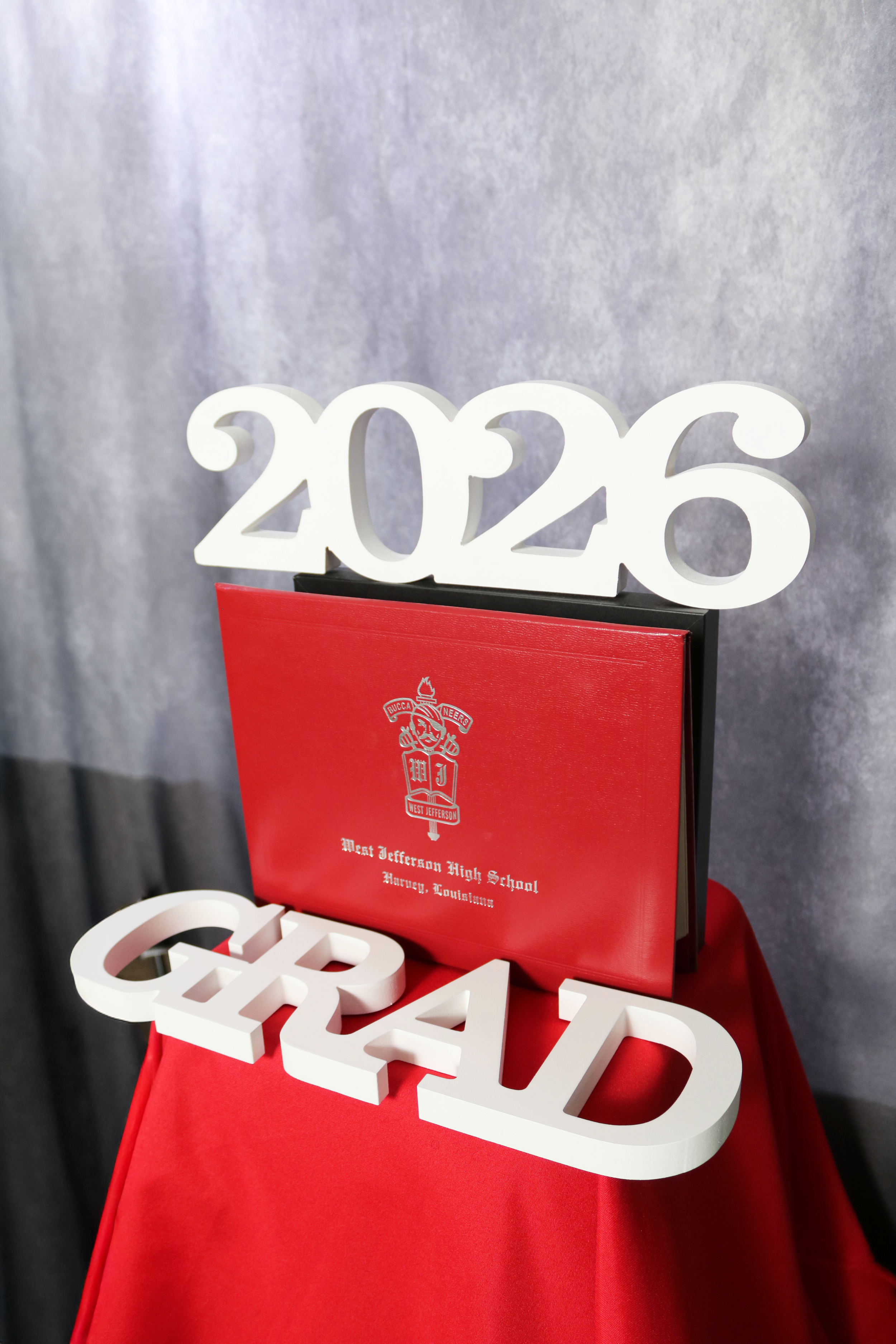 White 3D numbers spelling '2026' on a black stand behind a red plaque with white text and logo for West Jefferson High School, with a gray curtain backdrop.