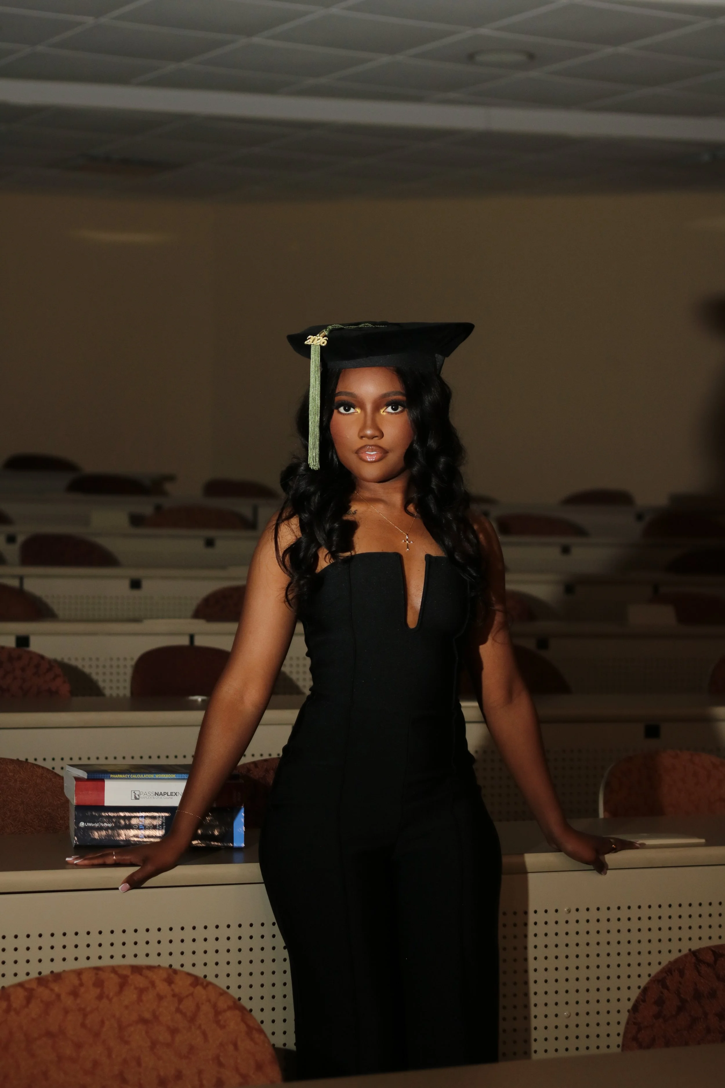 A woman with long, wavy black hair and gold makeup, wearing a black academic gown and cap, is standing in a lecture hall with empty chairs and desks, posing with one hand on a desk.