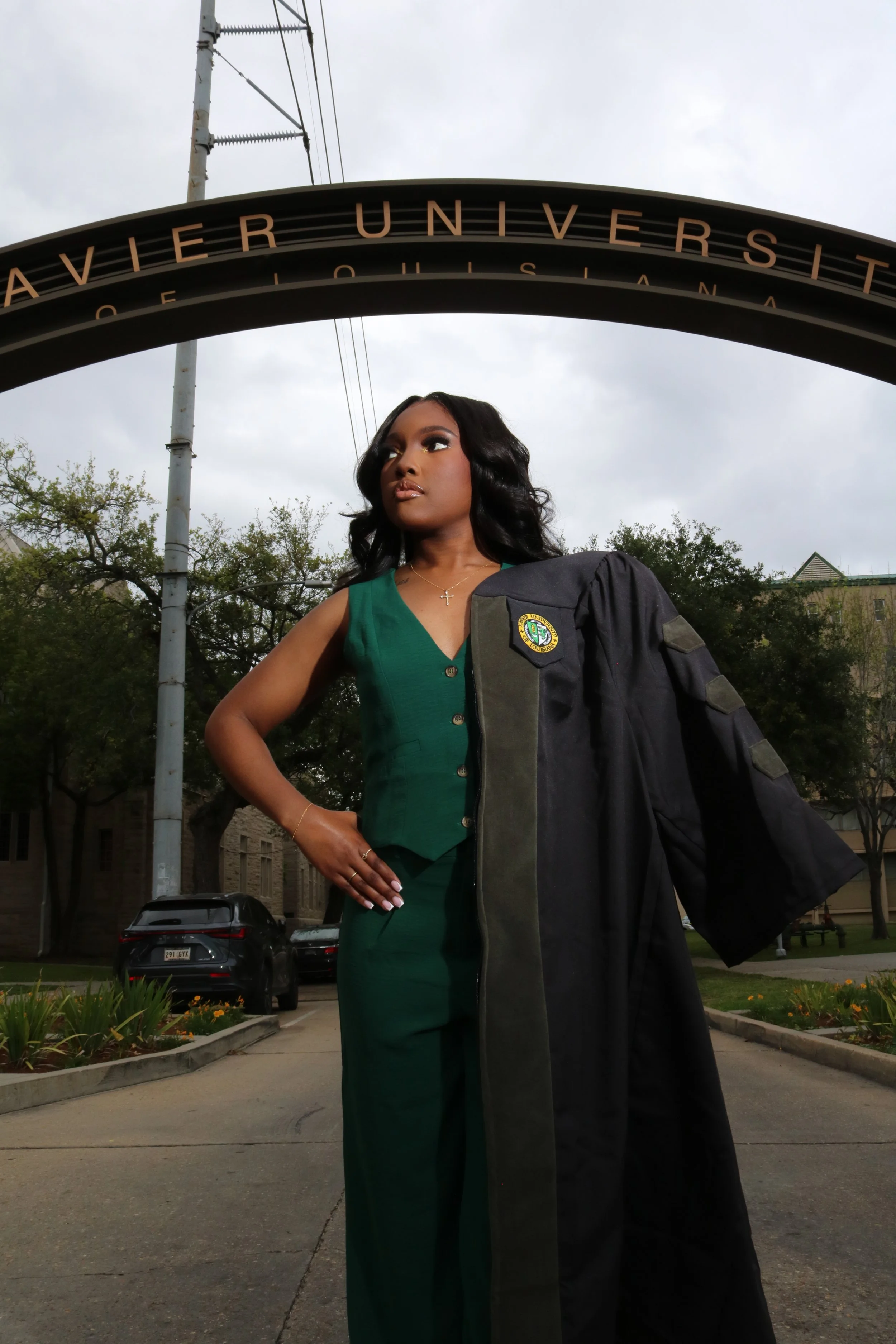 A woman wearing a green dress and holding a black graduation gown stands outdoors under an archway that reads "AUBER UNIVERSITY". There are trees, buildings, and parked cars in the background.