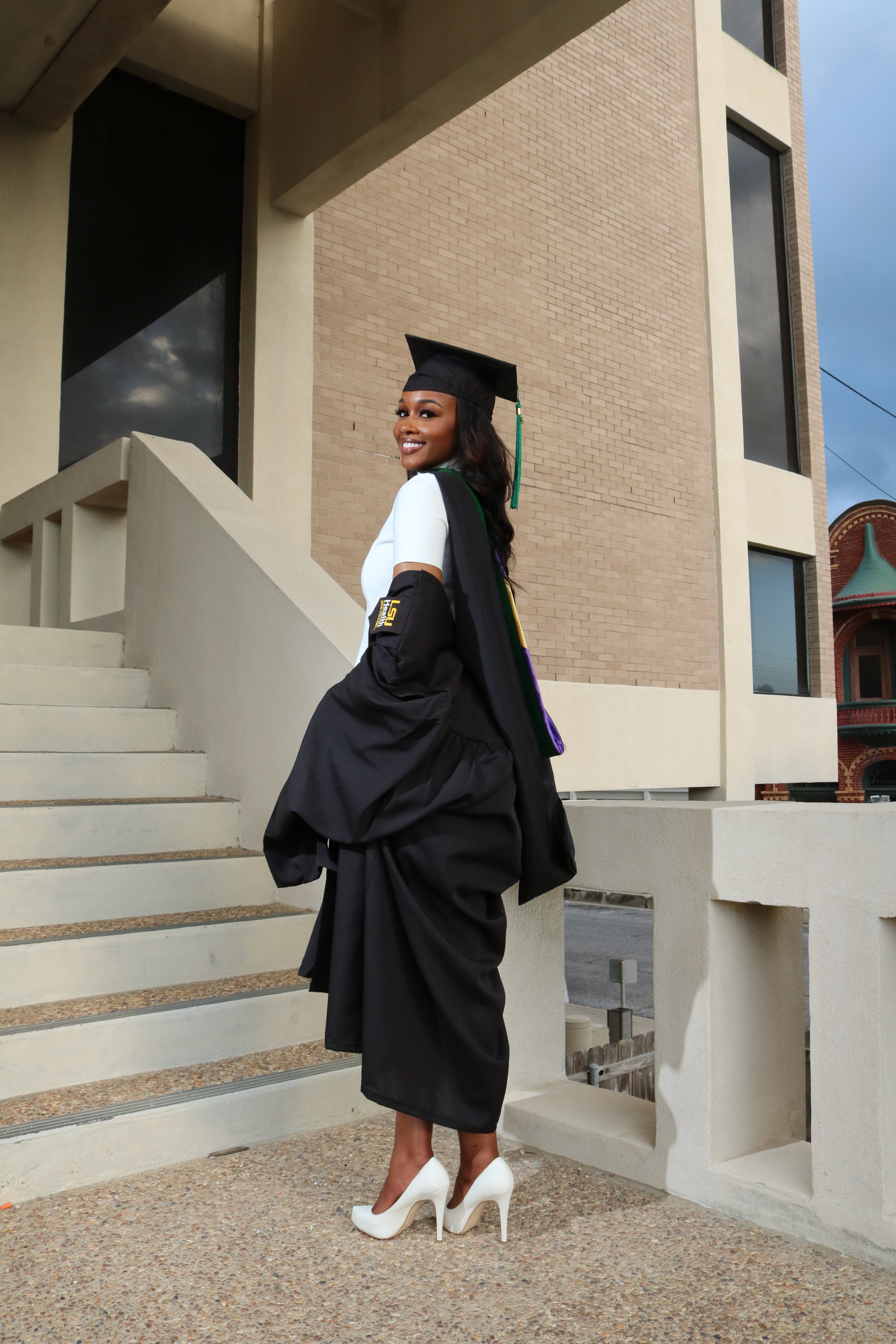 A woman in a graduation cap and gown, wearing white high heels, is standing outside on concrete stairs in front of a modern building with large windows and brick walls.