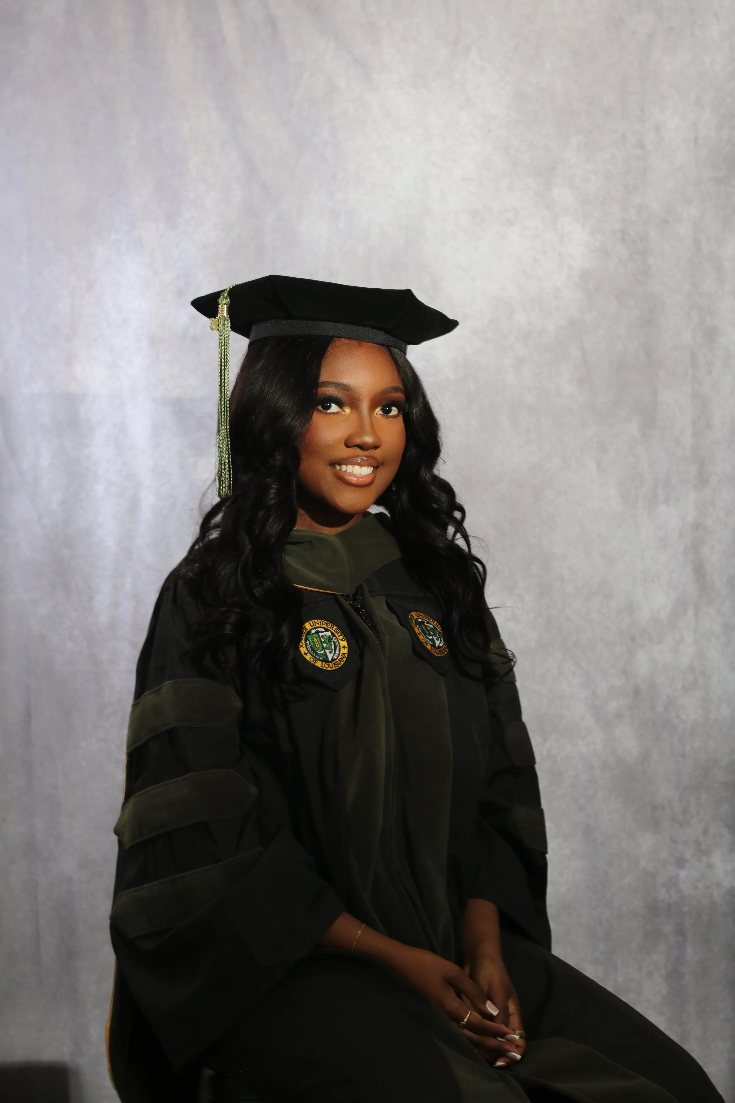 A woman in a graduation cap and gown sitting against a gray background.