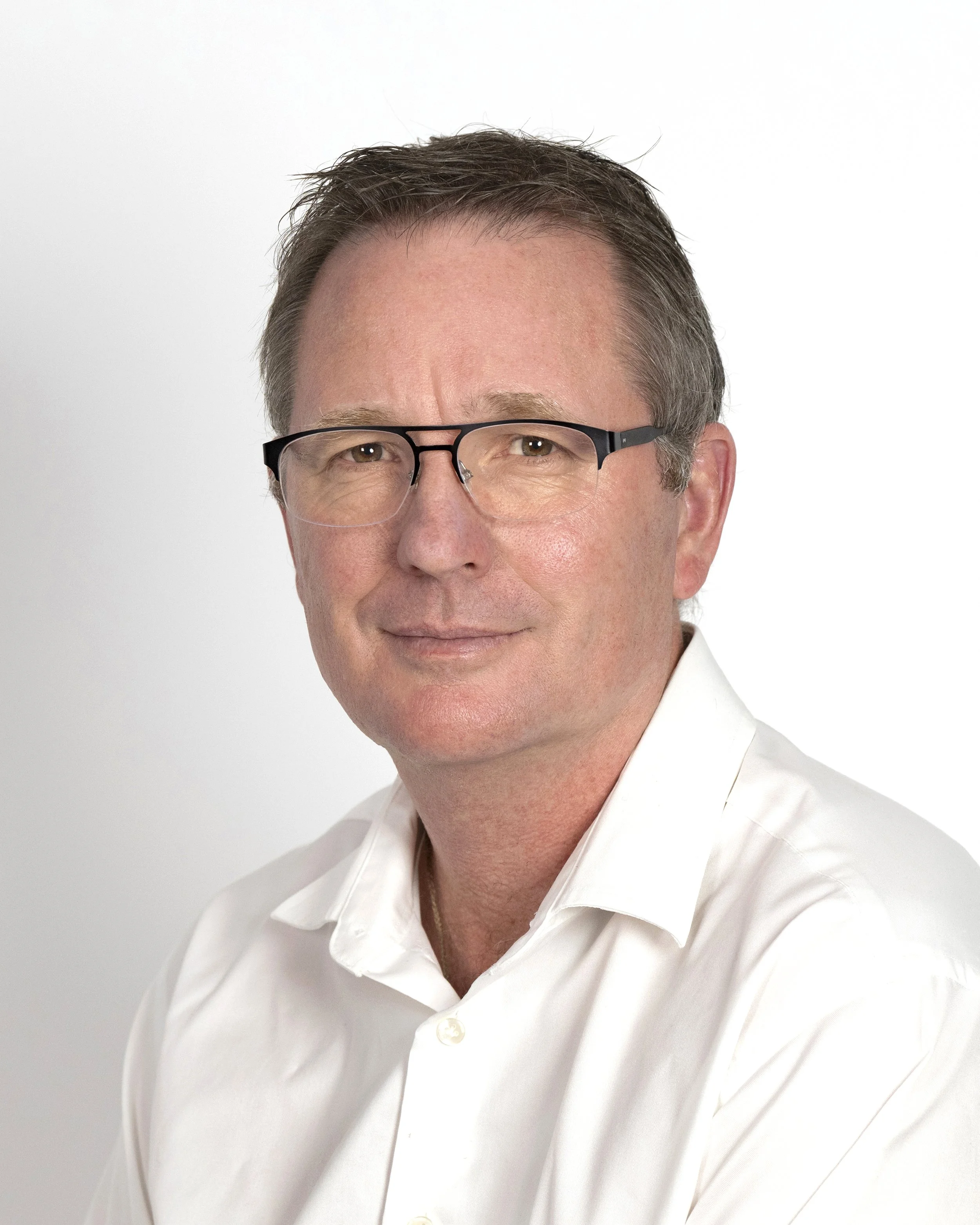 Headshot of a middle-aged man with short brown hair, glasses, and wearing a white collared shirt, posed against a plain white background.