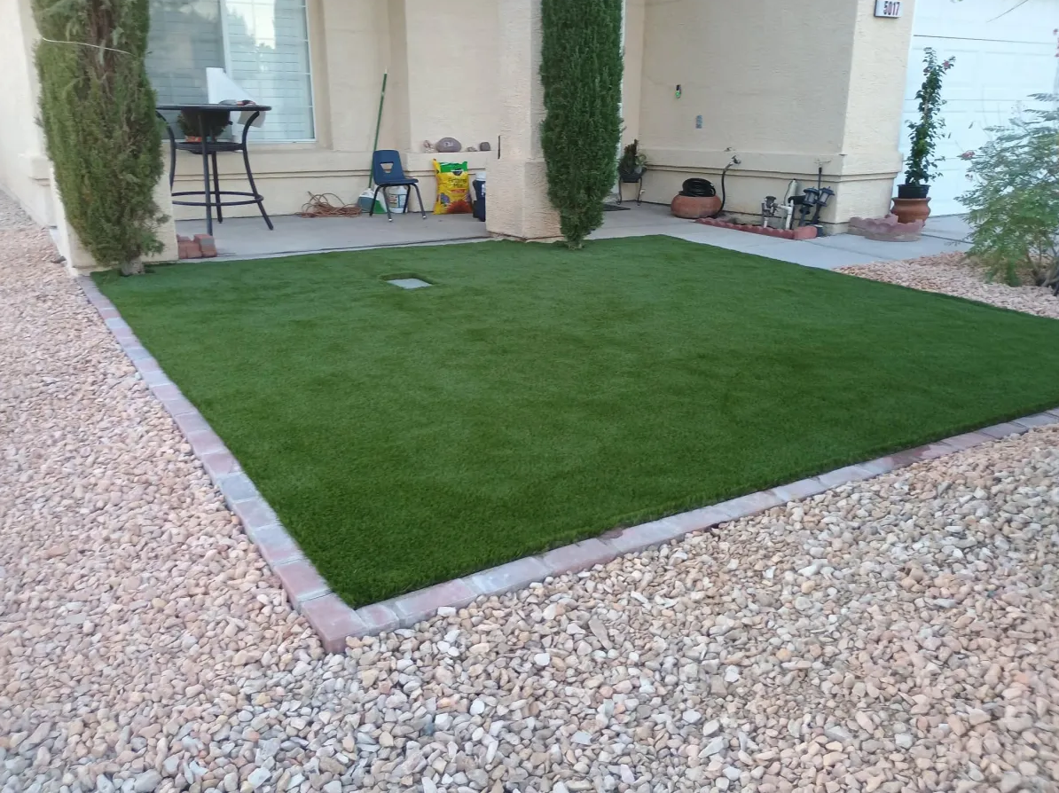 A backyard with a small, neatly edged artificial grass lawn surrounded by gravel and bordered with bricks, with a house wall and patio furniture in the background.