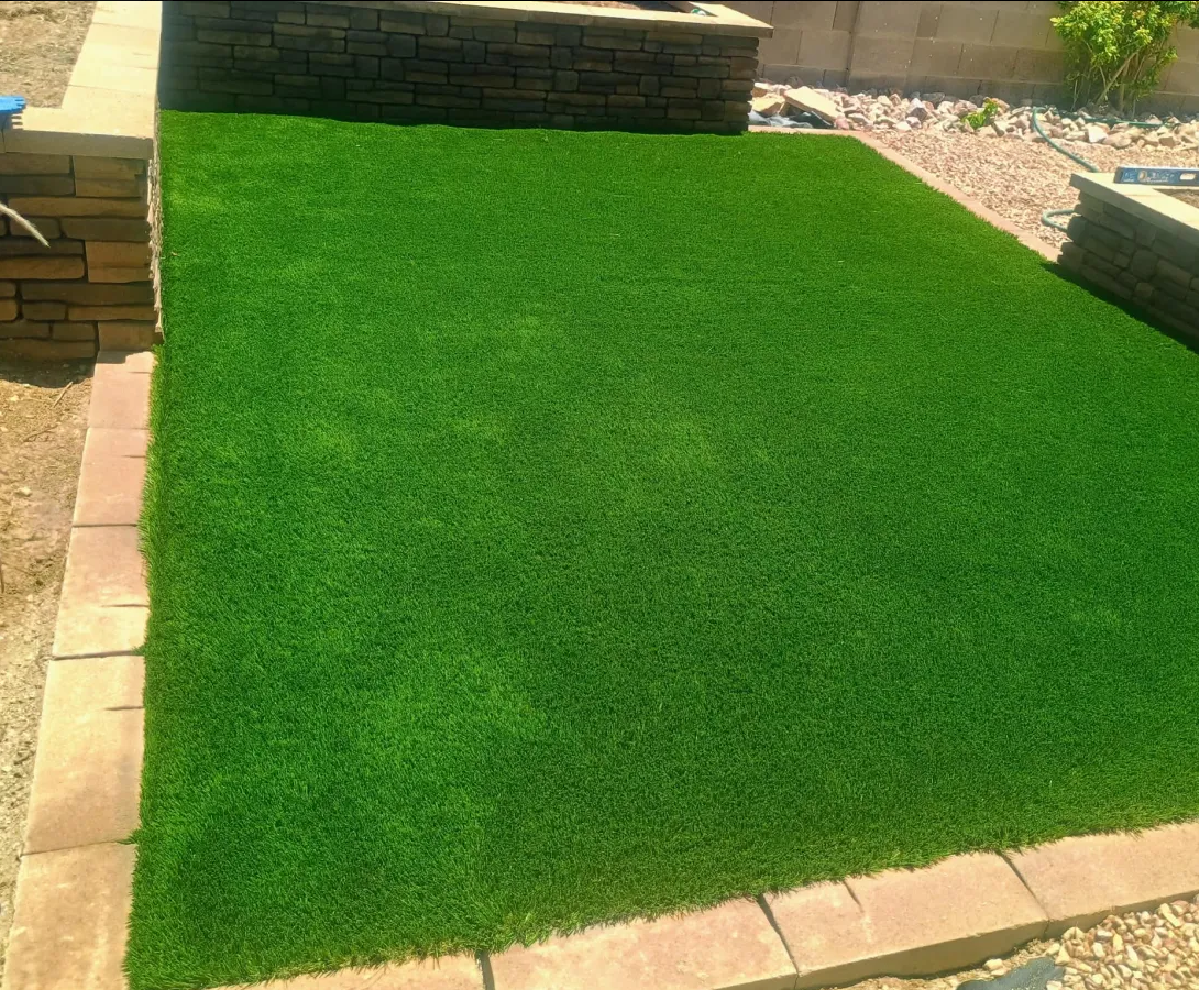 Artificial grass installed in a landscaped backyard area, bordered by stone pavers.