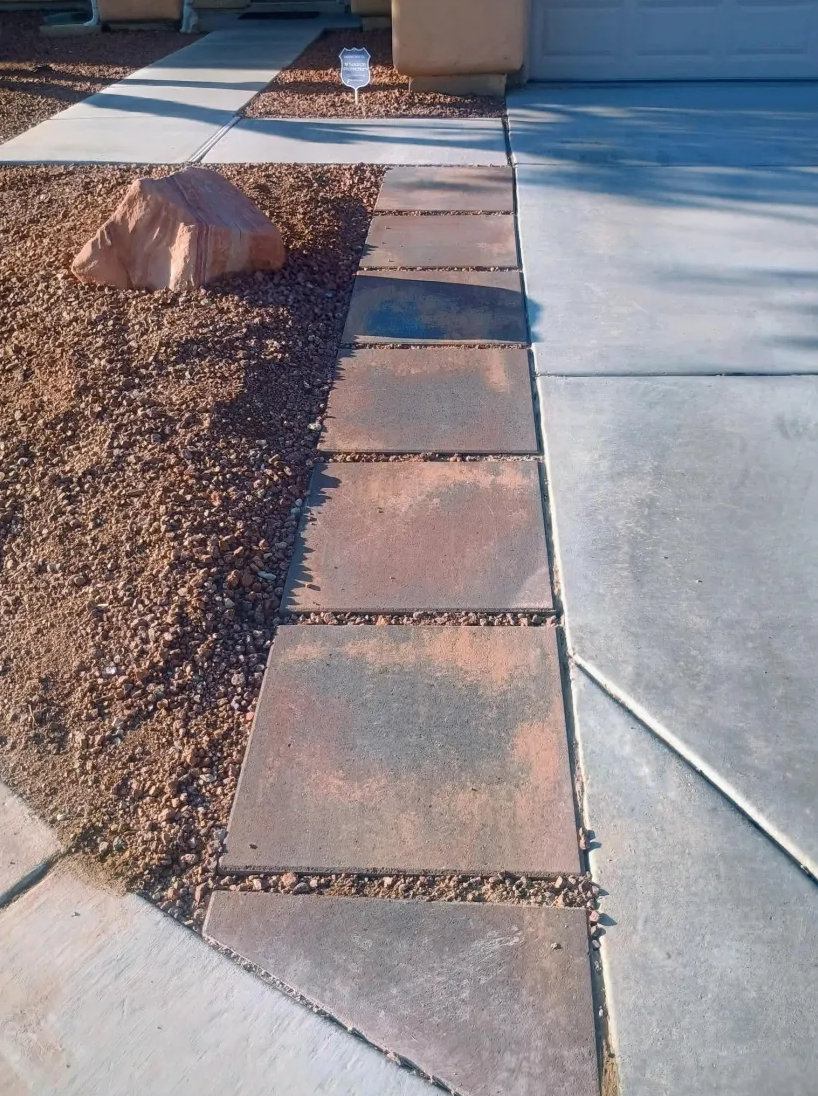A walkway with a large rock on dirt and gravel, leading to a sidewalk and a garage with a small sign in the background.