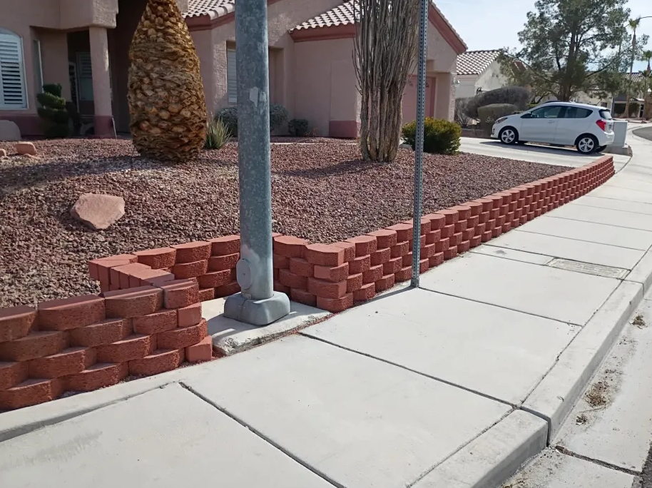 A new brick wall border along the sidewalk in front of a house with desert landscaping, including a large cactus and small bushes, and a parked white car in the background.