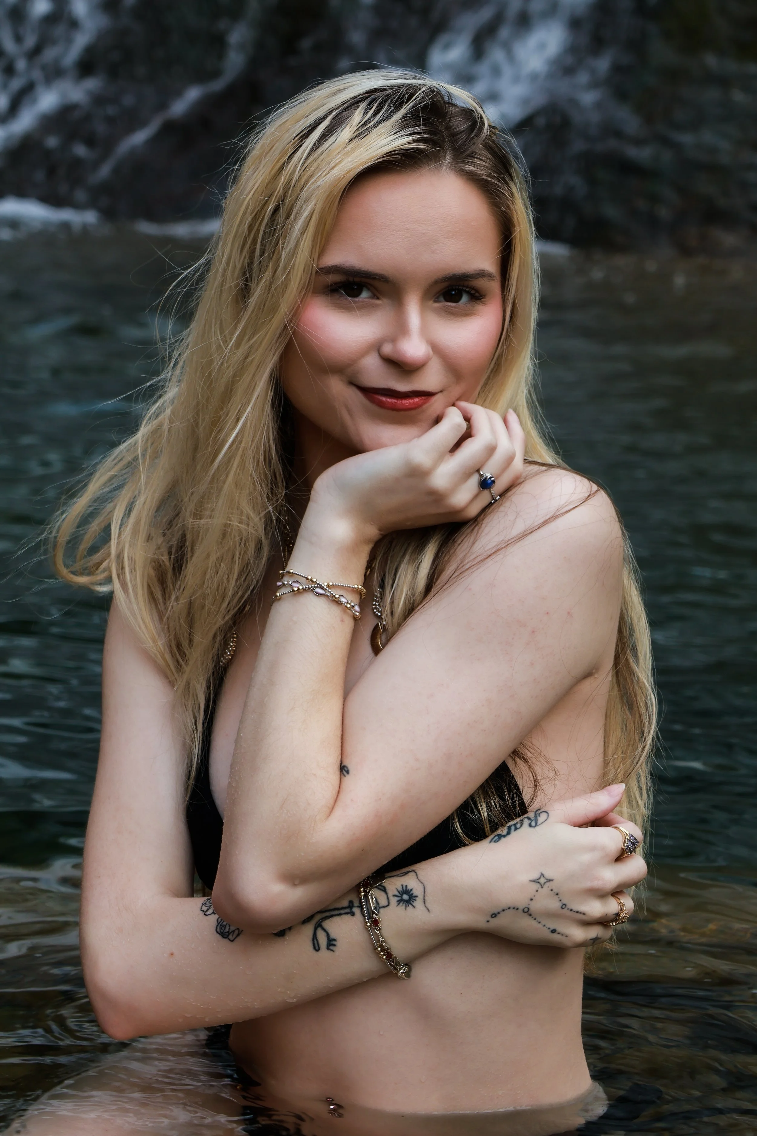 A young woman with long blonde hair posing in a body of water with rocks in the background.