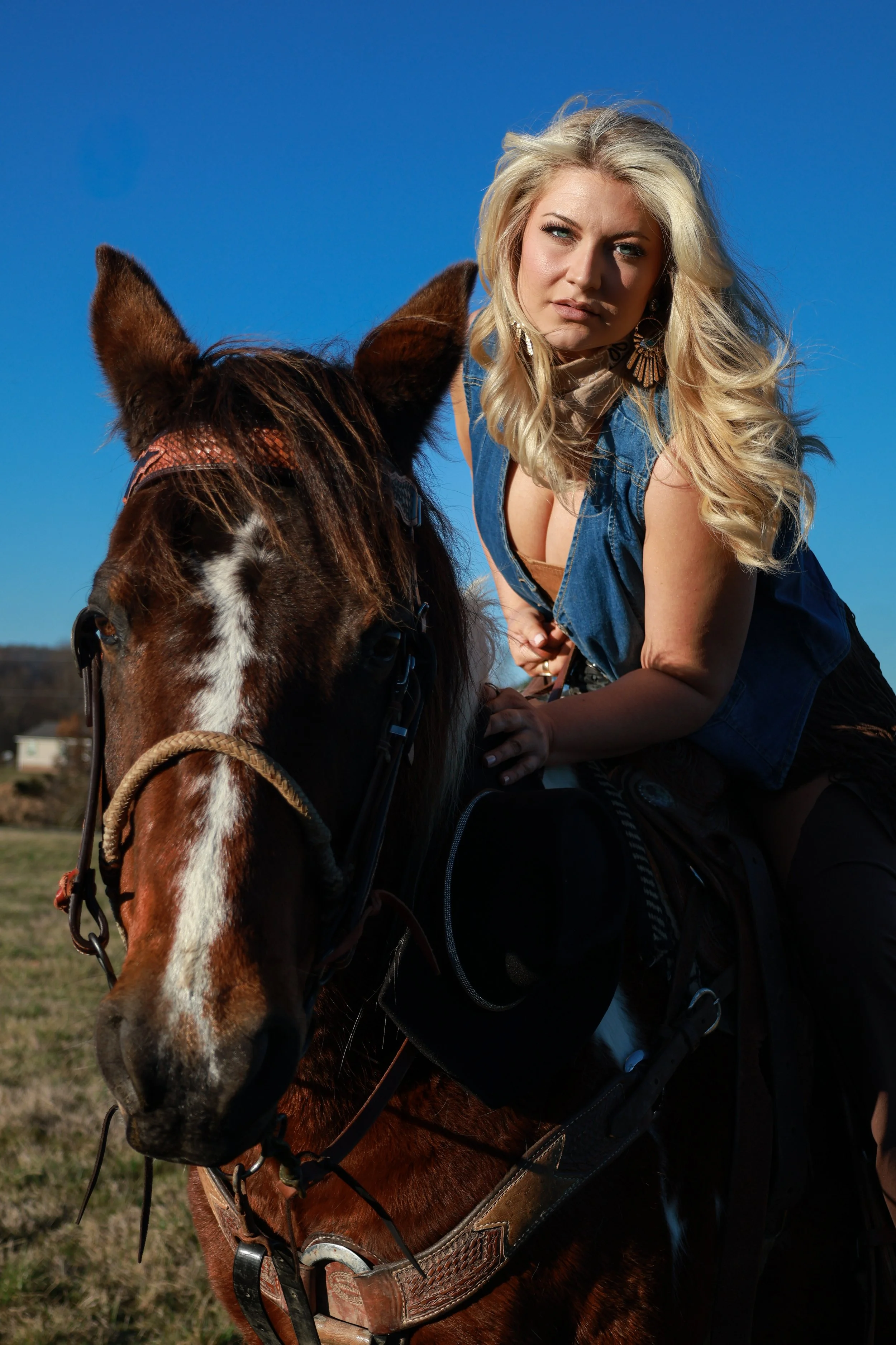 A blonde woman in a sleeveless denim vest riding a brown and white horse outdoors on a clear day.