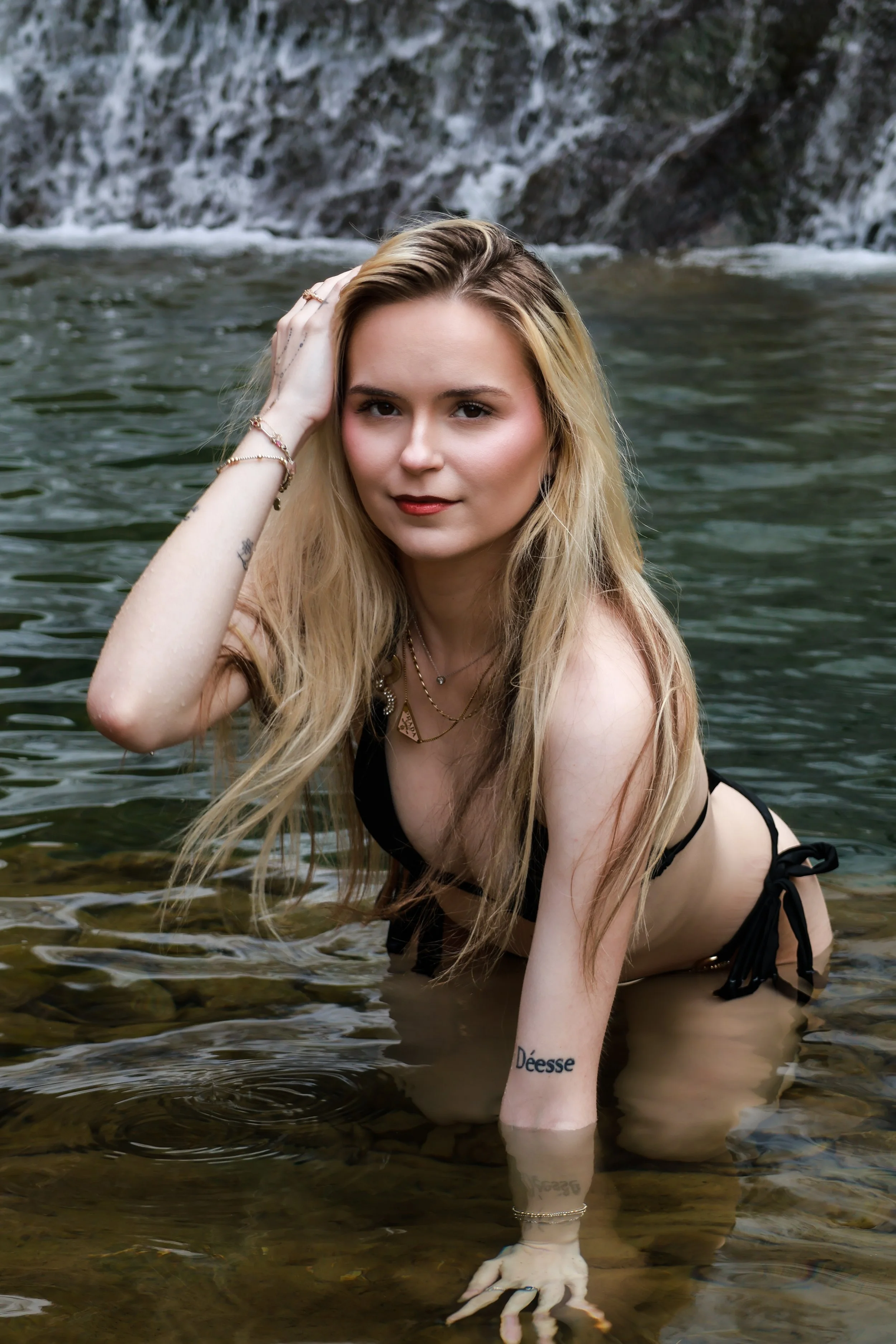 A young woman with long blonde hair in a black bikini is kneeling in a natural body of water with a rocky background.