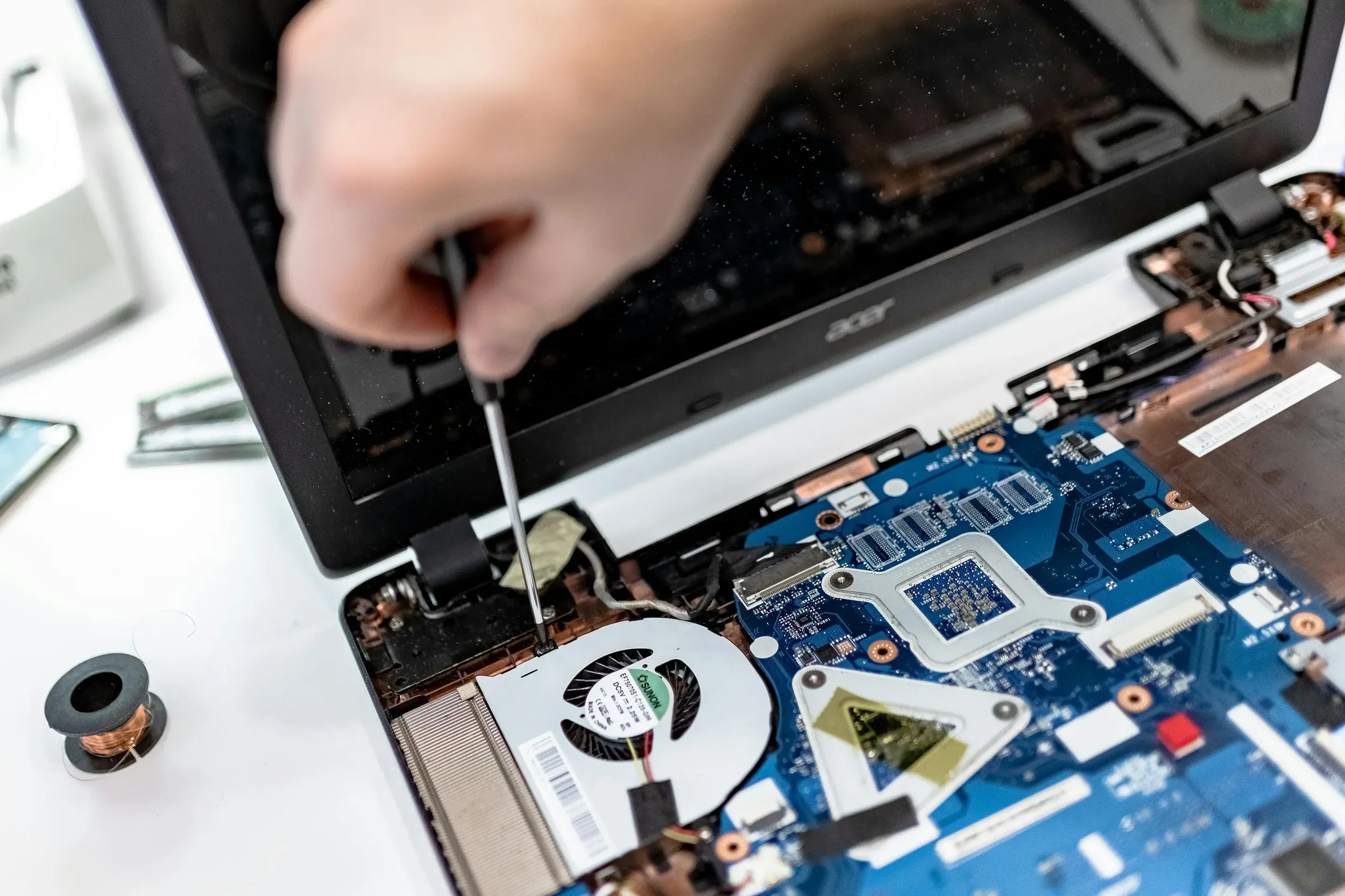 A person using a screwdriver to repair or upgrade a laptop, with the back cover removed to expose the internal components including the computer's motherboard and cooling fan.