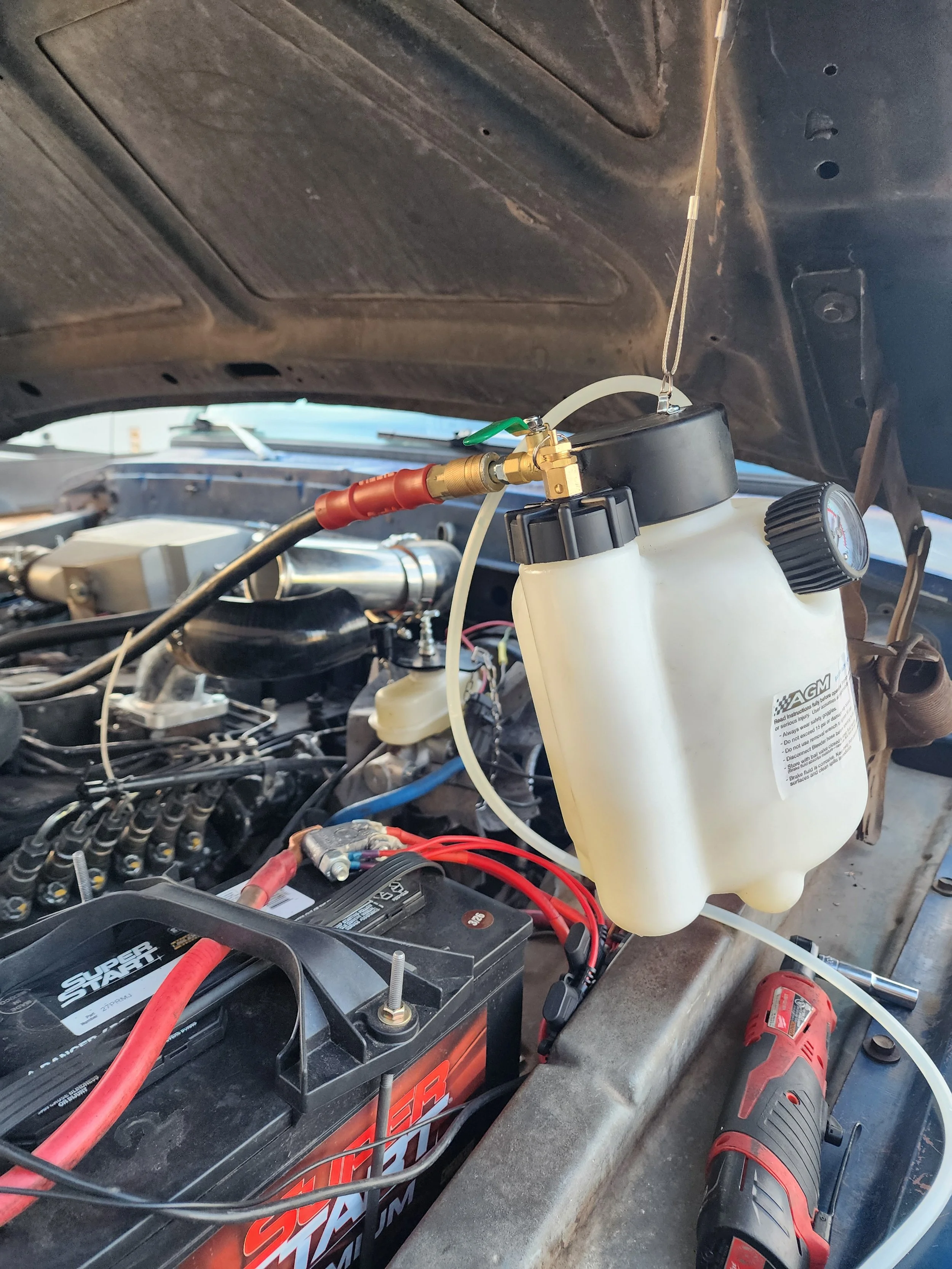 The image shows the interior of a car with the hood open, revealing a near-finished radiator coolant system. In the foreground, there's a car battery with red and black cables connected, and a cordless drill resting on the surface. A white coolant reservoir with a black cap and a pressure gauge is mounted near the engine bay. Various wires, hoses, and engine components are visible in the background.