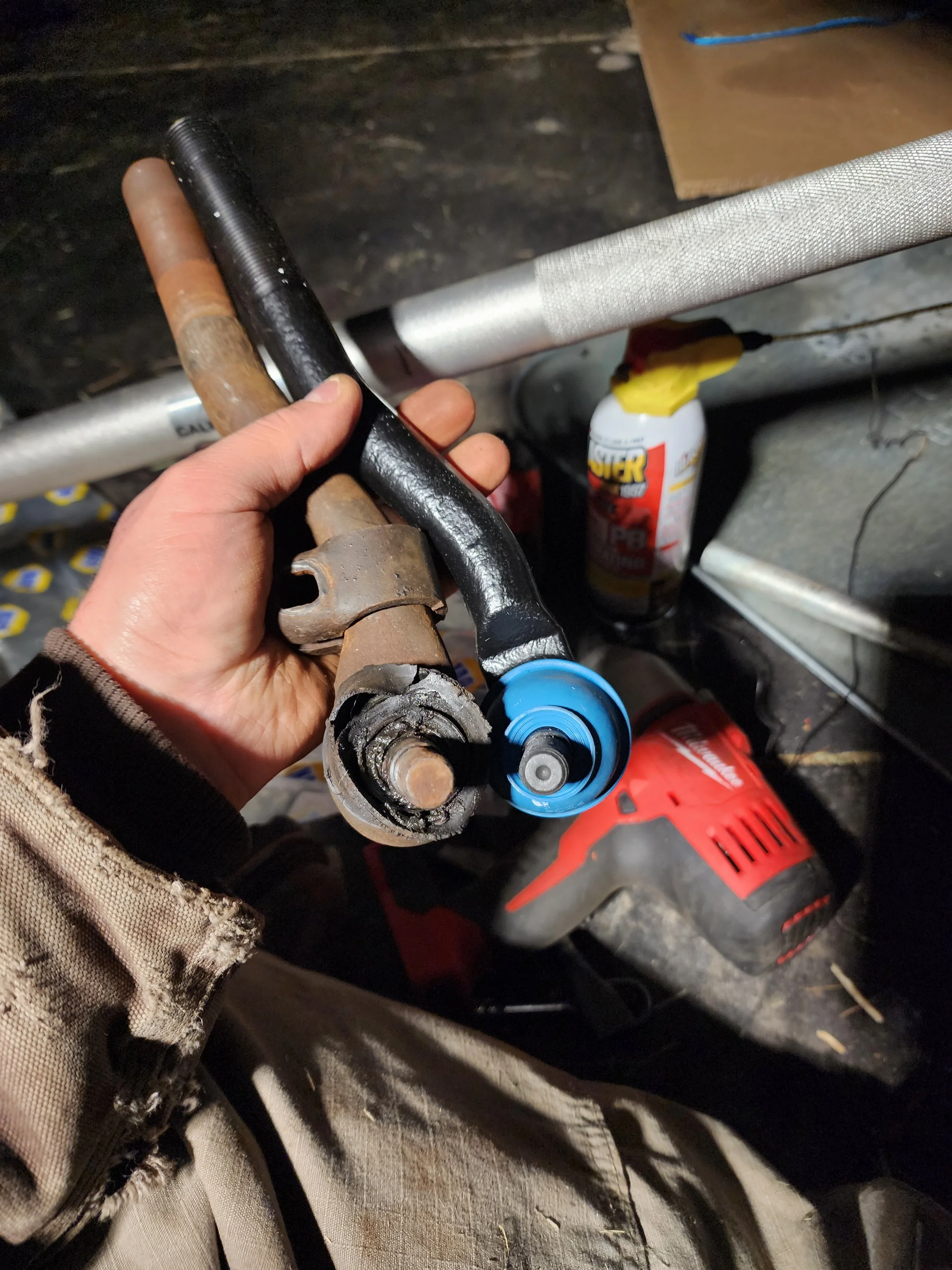 A person holding a worn-out tie rod end in a garage workshop. Various tools and other equipment are visible in the background.