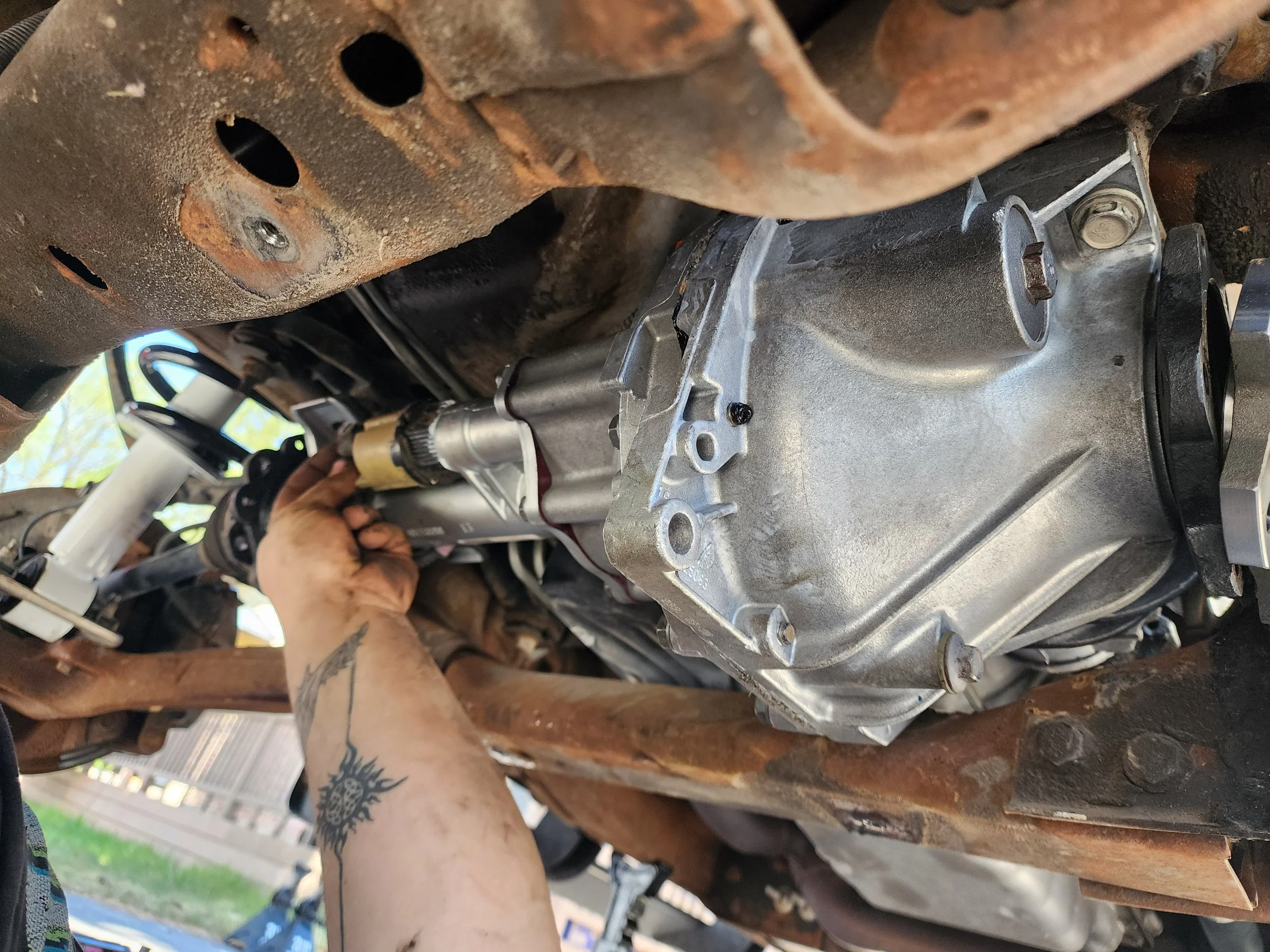 A person working underneath a vehicle, installing  a front differential, surrounded by rusted car parts.