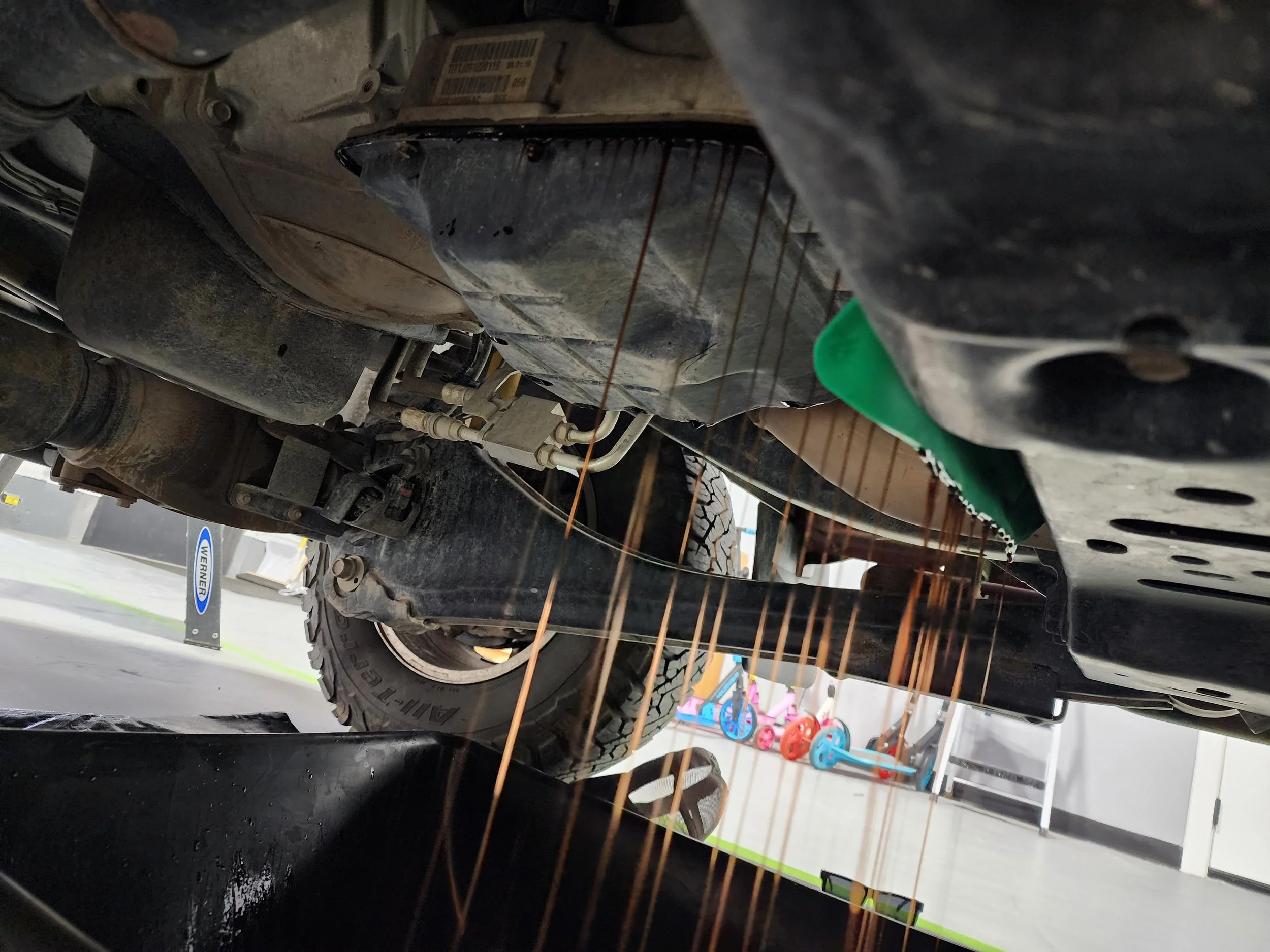 Underneath view of a vehicle showing a transmission service and underside components in a garage with children's tricycles in the background.