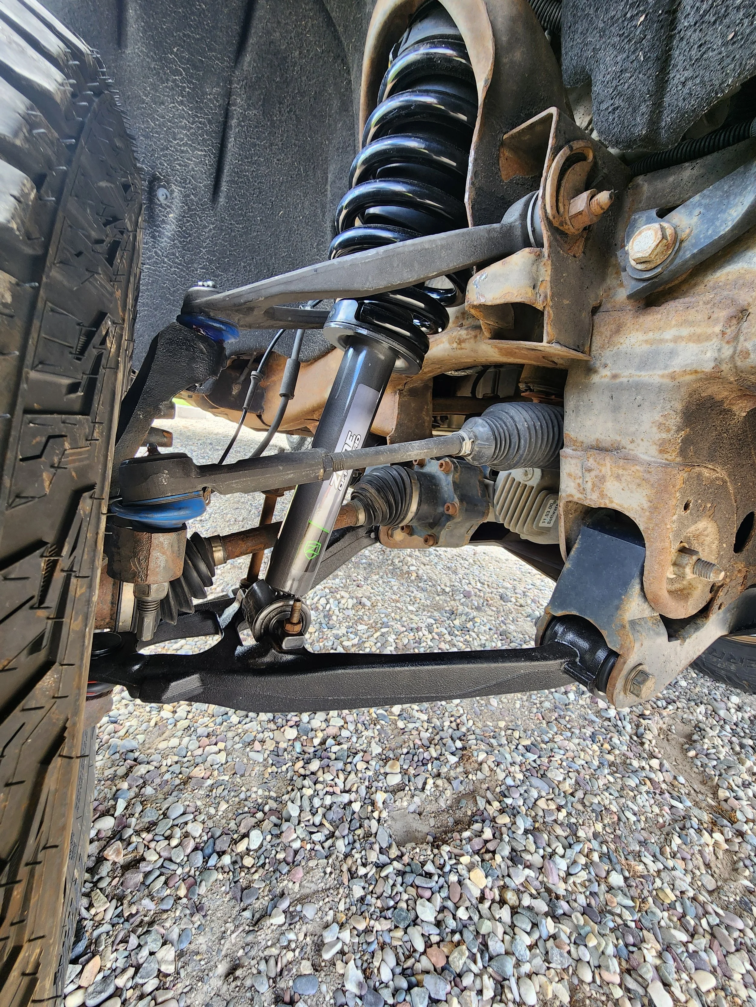 Close-up view of a vehicle's suspension system showing a coil spring, shock absorber, and various suspension components, with the vehicle's wheel and tire visible on the left side, and gravel ground below.
