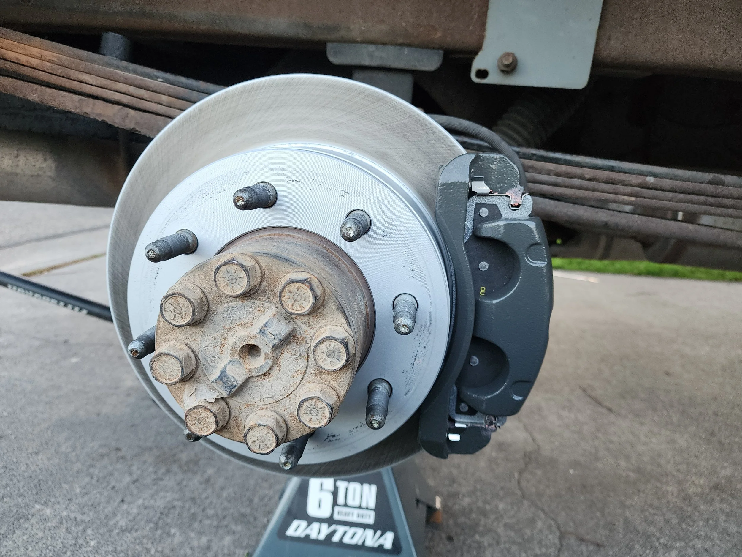 Close-up of a vehicle's disc brake assembly, showing the brake rotor and wheel hub with lug studs, with a black and white jack stand '6 TON DAYTONA' visible below.