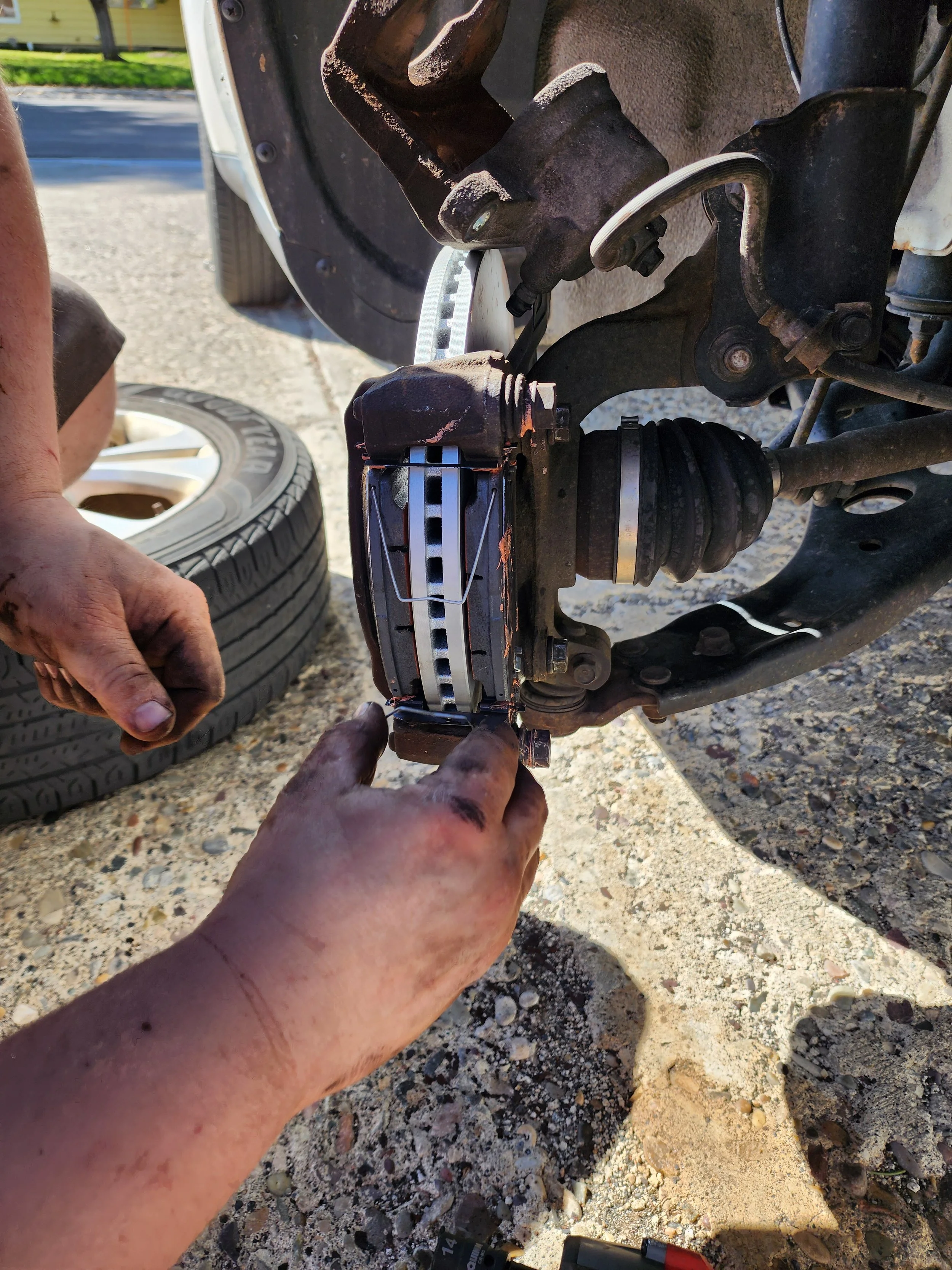 Hands working on a vehicle's suspension system, removing or installing a brake rotor with tools, with a tire and gravel ground in an outdoor setting.